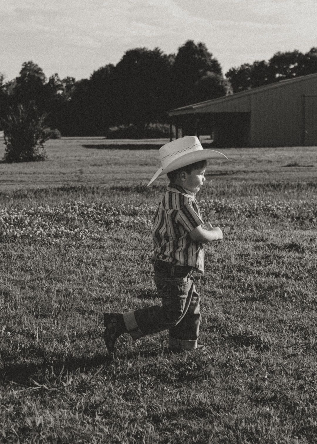 Black and white photo of a young boy in cowboy hat and boots running across a grassy field near a barn.