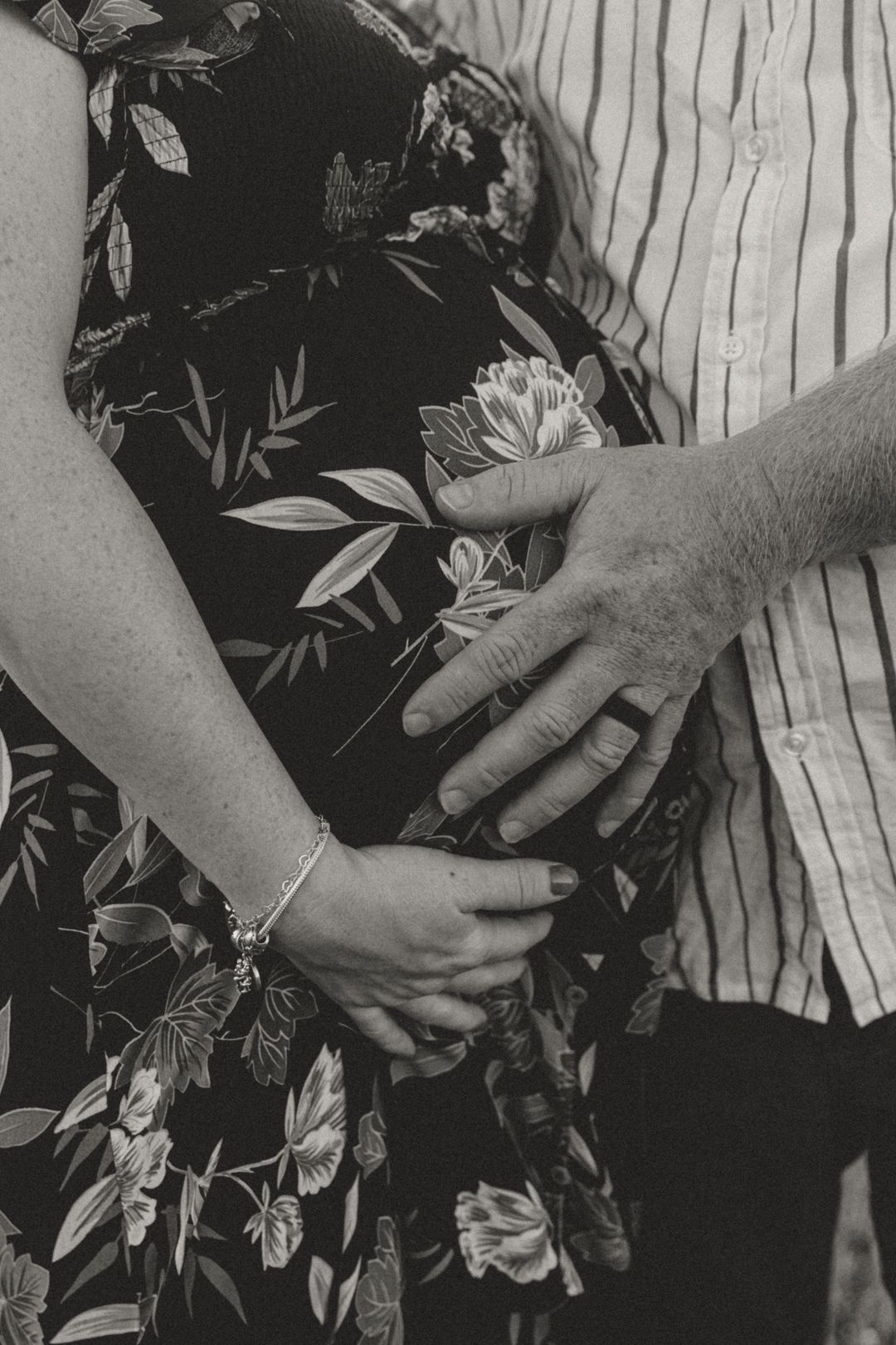 Black and white maternity photo of couple holding hands over pregnant belly in floral dress outdoors.