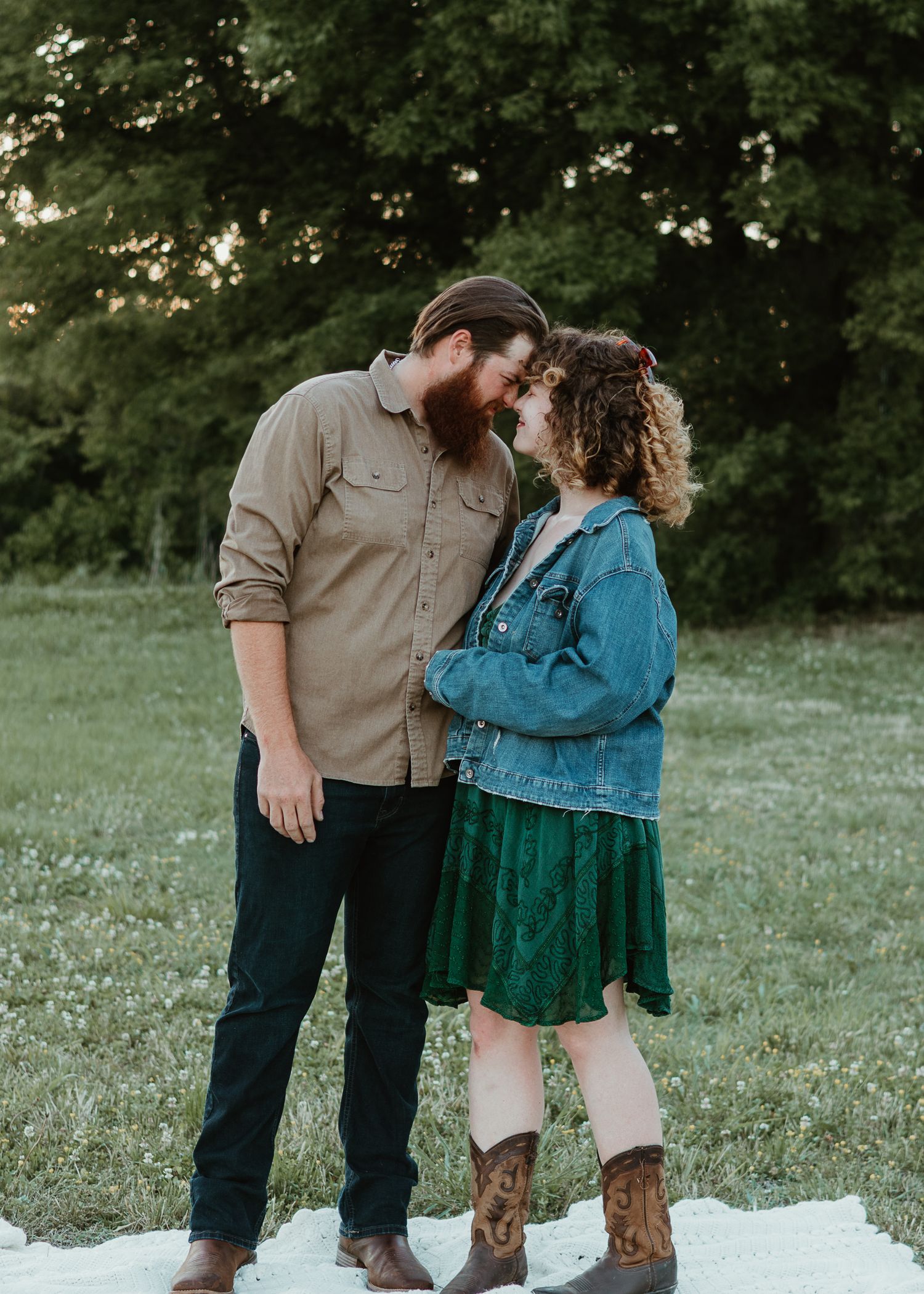 Couple sharing a romantic moment in a green field, man in tan shirt and woman in denim jacket and green dress.