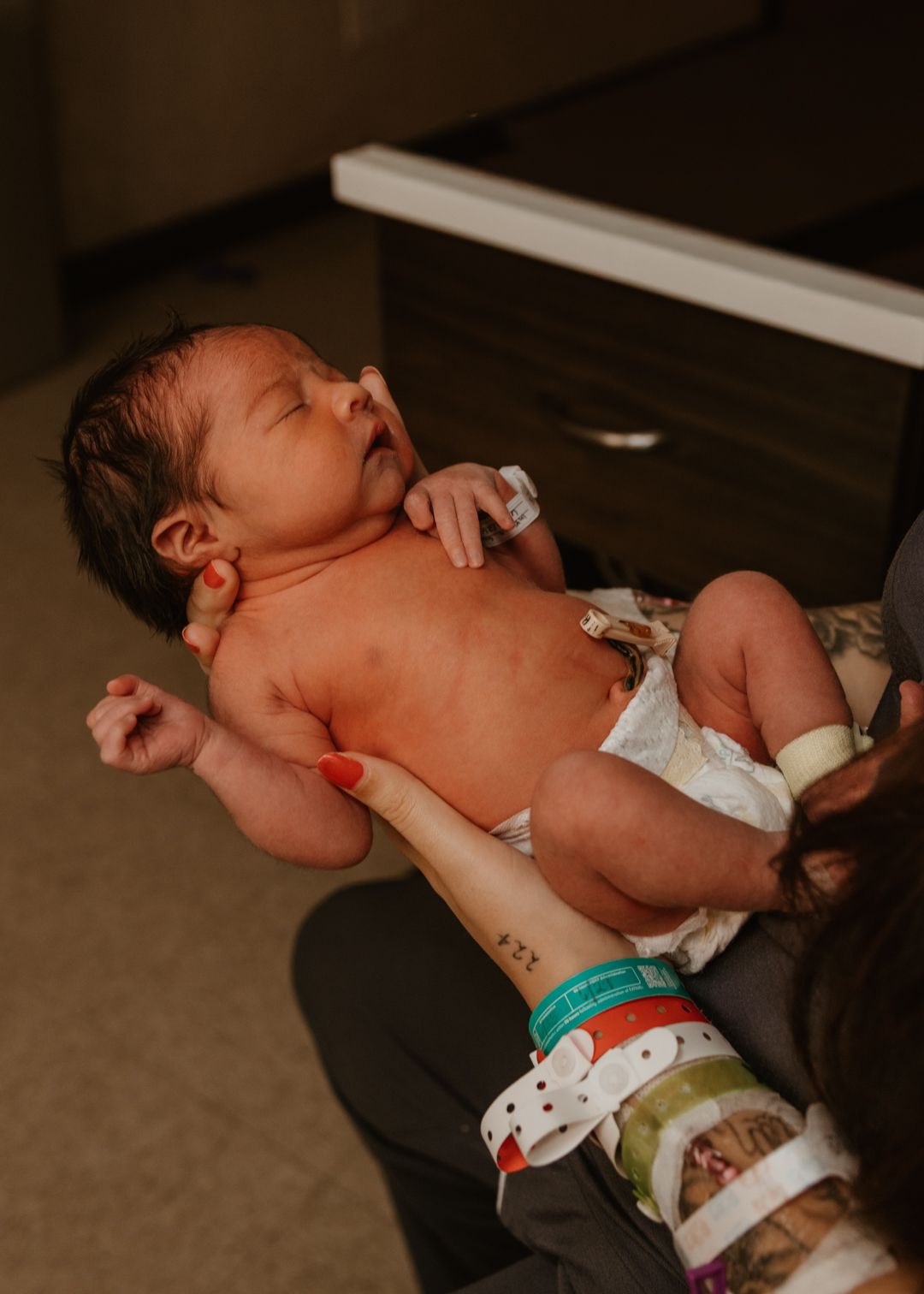 Newborn baby resting in parent's arms wearing a diaper with hospital ID bracelets visible.