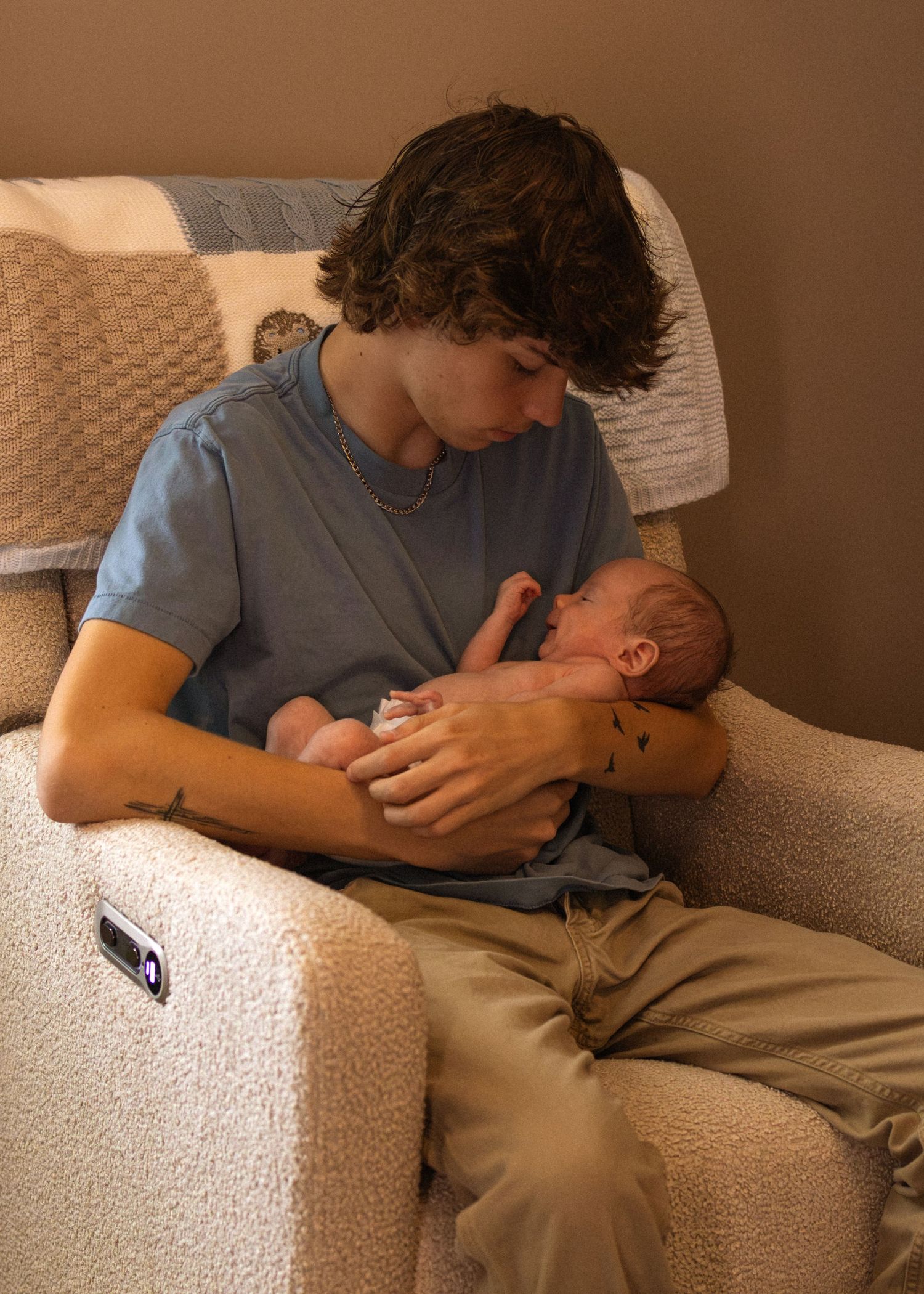 Young teenage boy with dark curly hair gently cradles a newborn baby in a recliner chair.