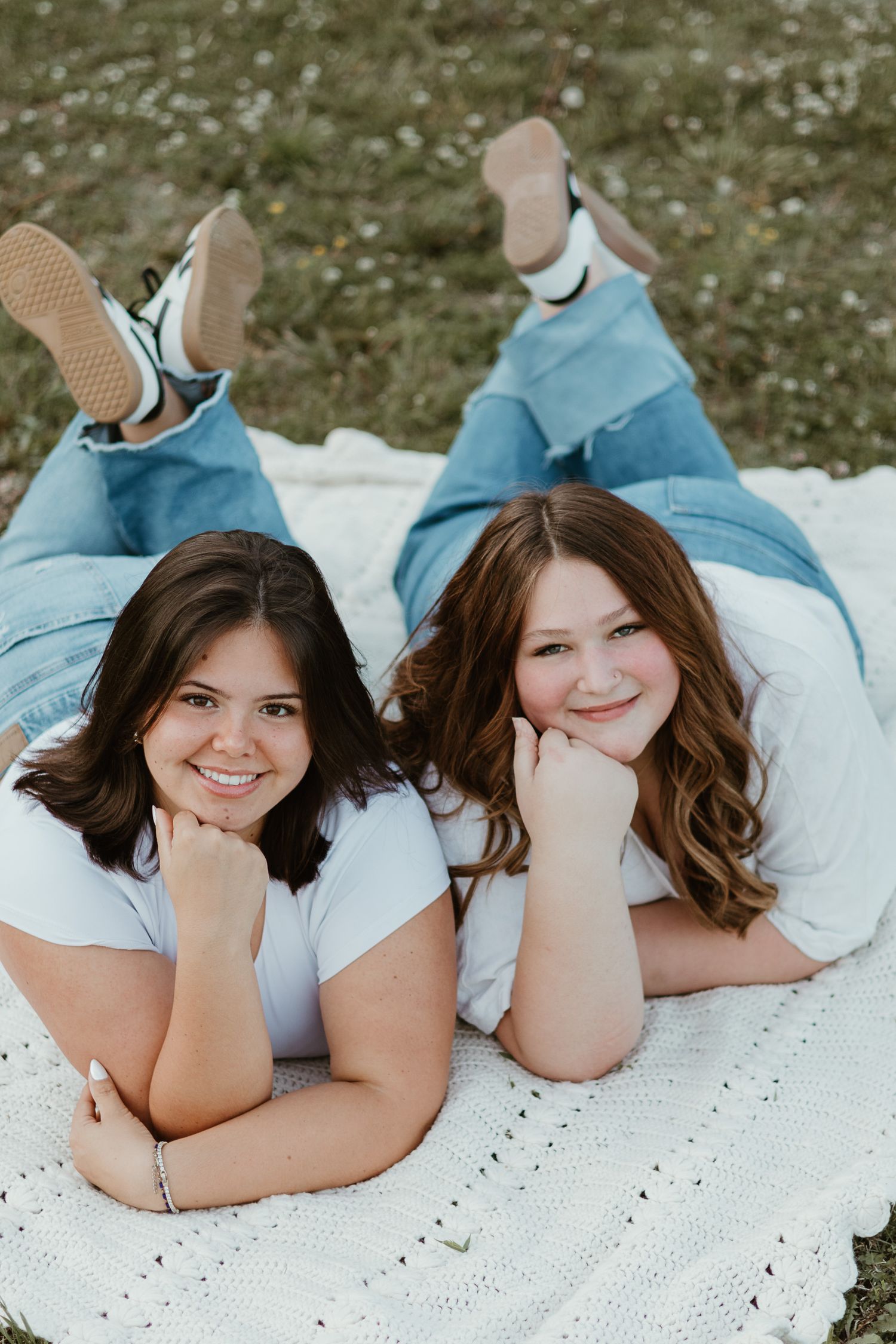 Two smiling young women in white tees and jeans lying on a white blanket outdoors, posing with chins resting on hands.