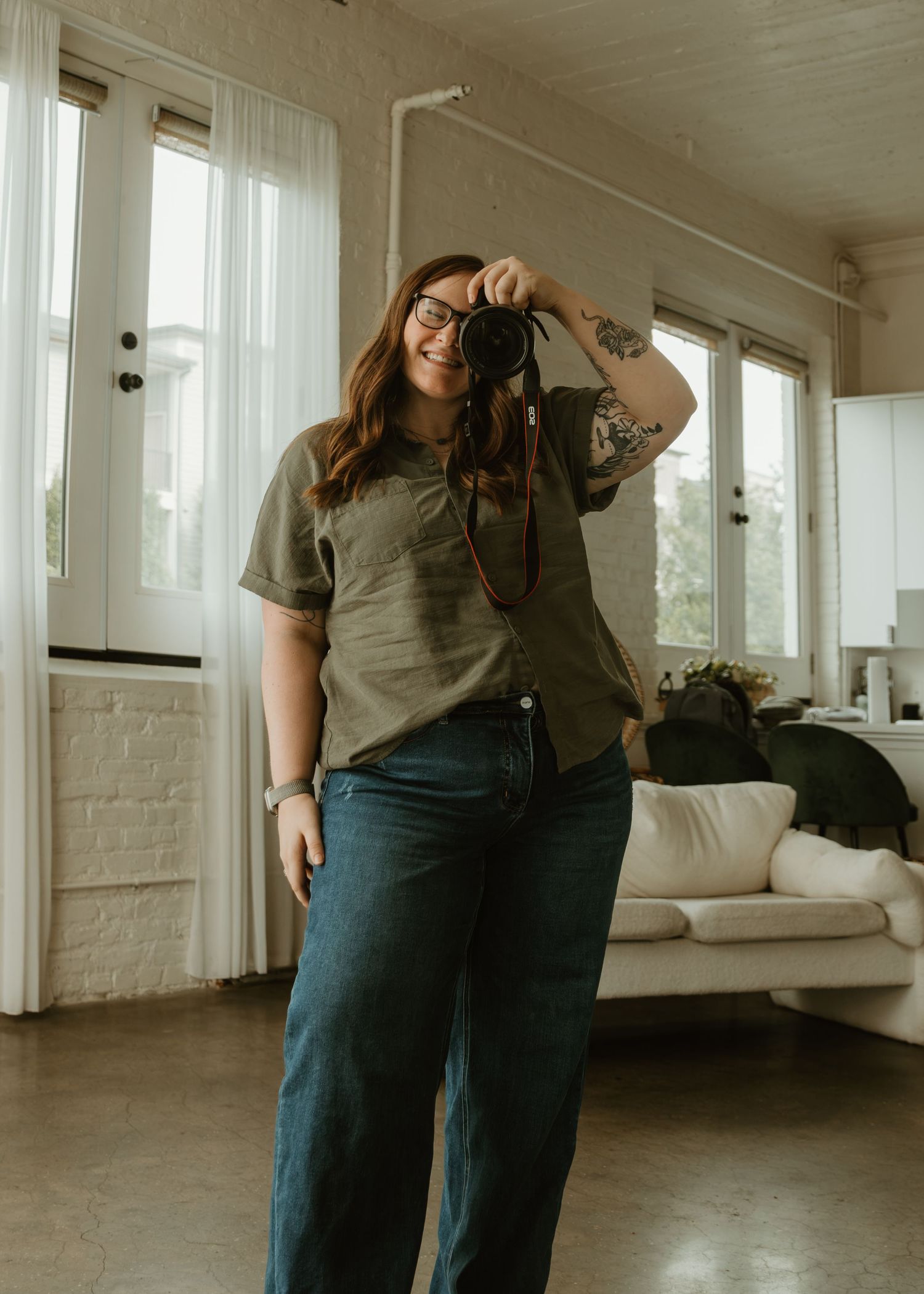 A smiling woman holds a camera up to her face while standing in a bright studio, wearing a green tee and wide-leg jeans.