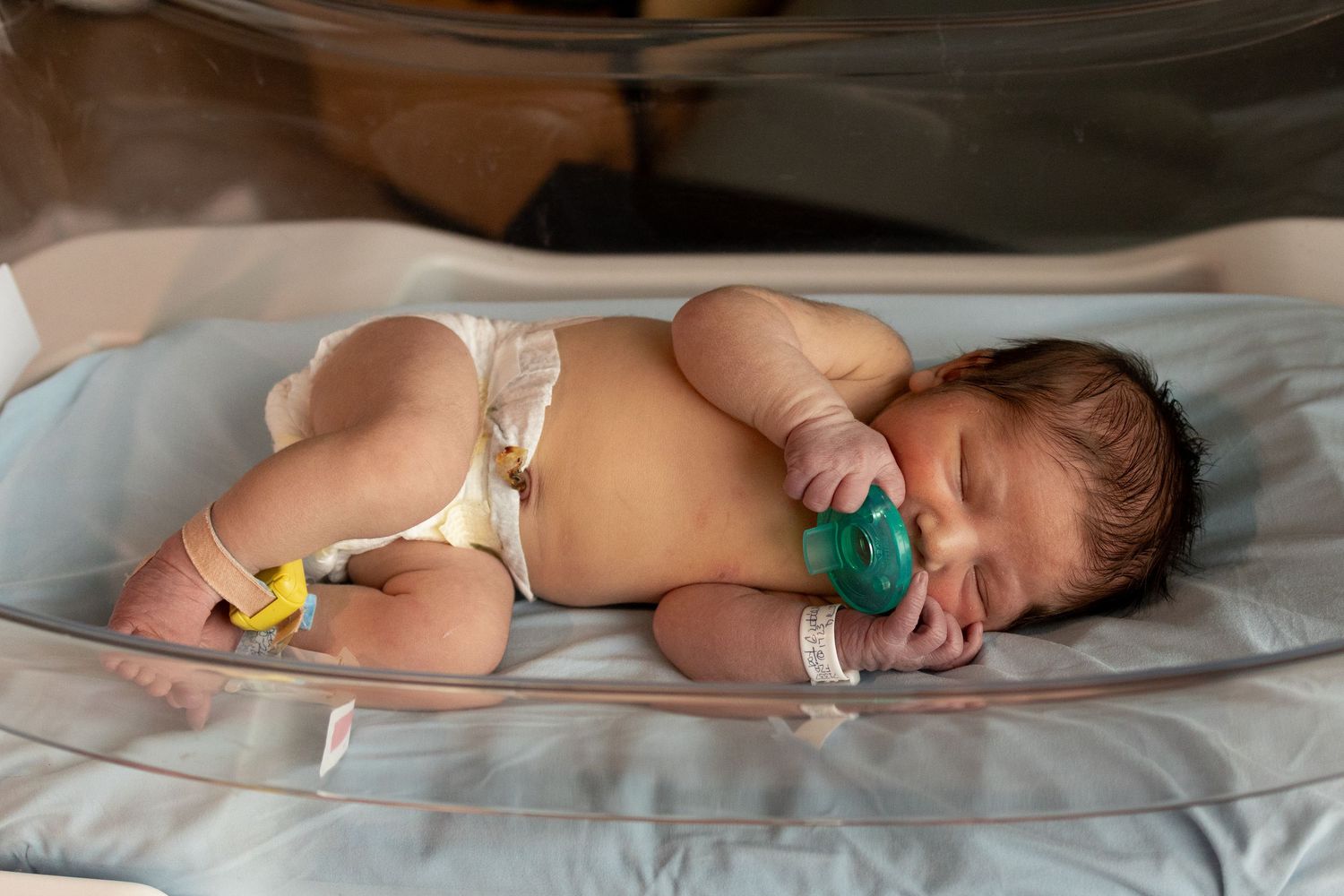 Newborn baby sleeping in hospital bassinet with pacifier and identification bracelets.
