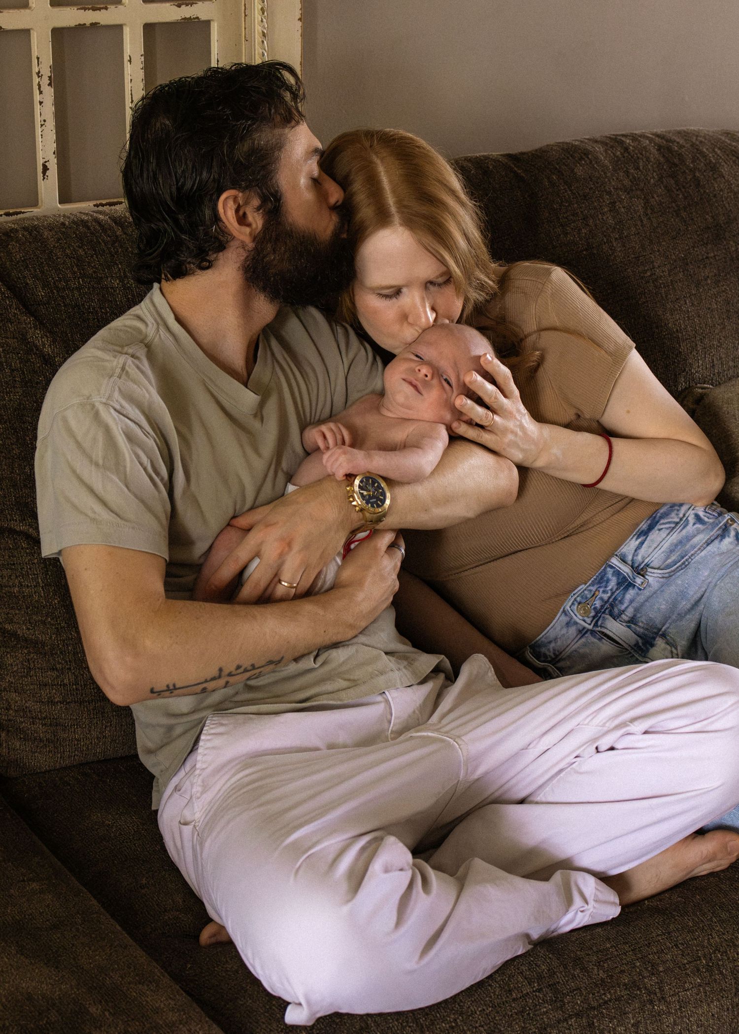 New parents sitting on a couch kissing their newborn baby, father wearing a gold watch and red bracelet.