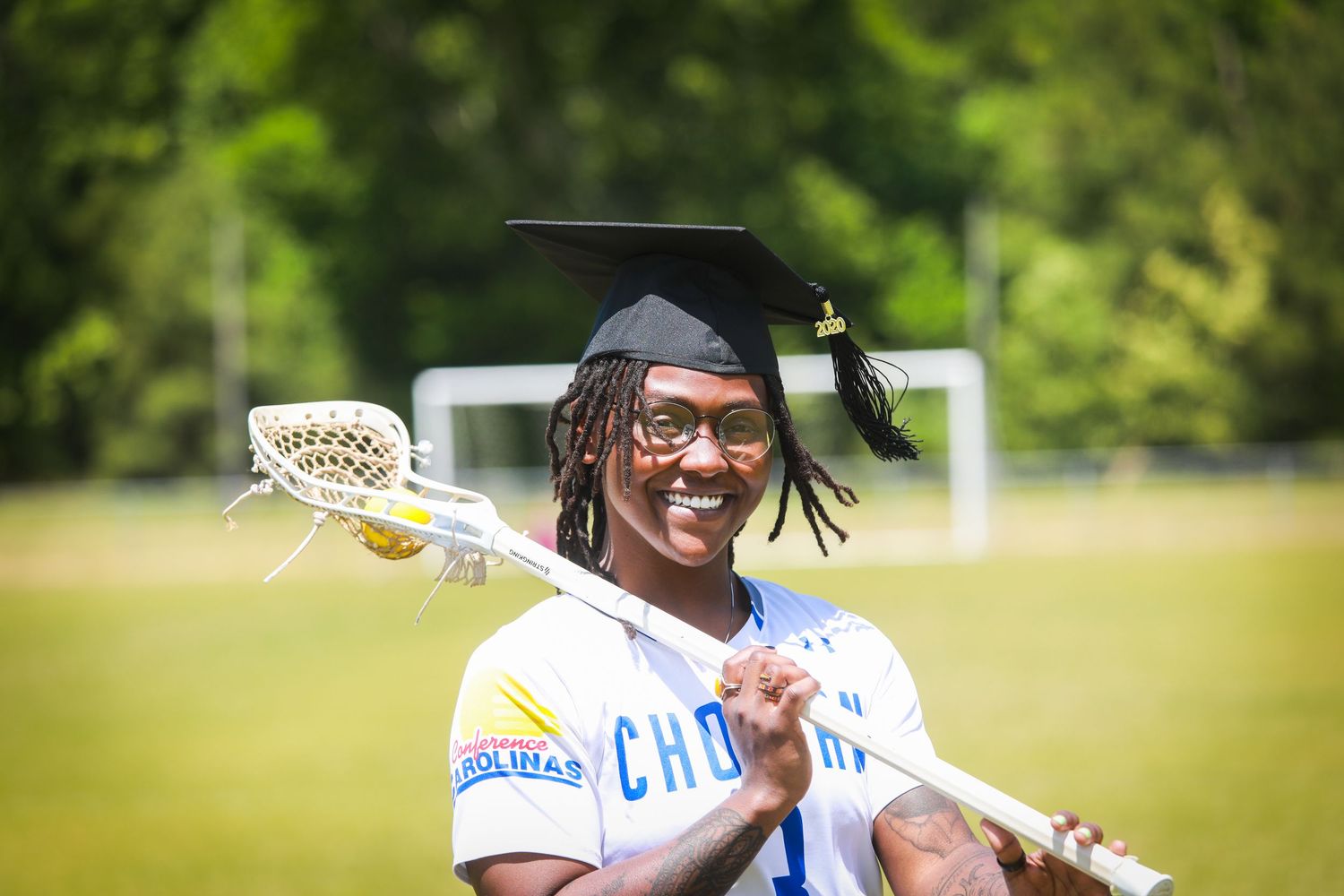 Smiling graduate in cap holding lacrosse stick on green field, wearing blue and white jersey.