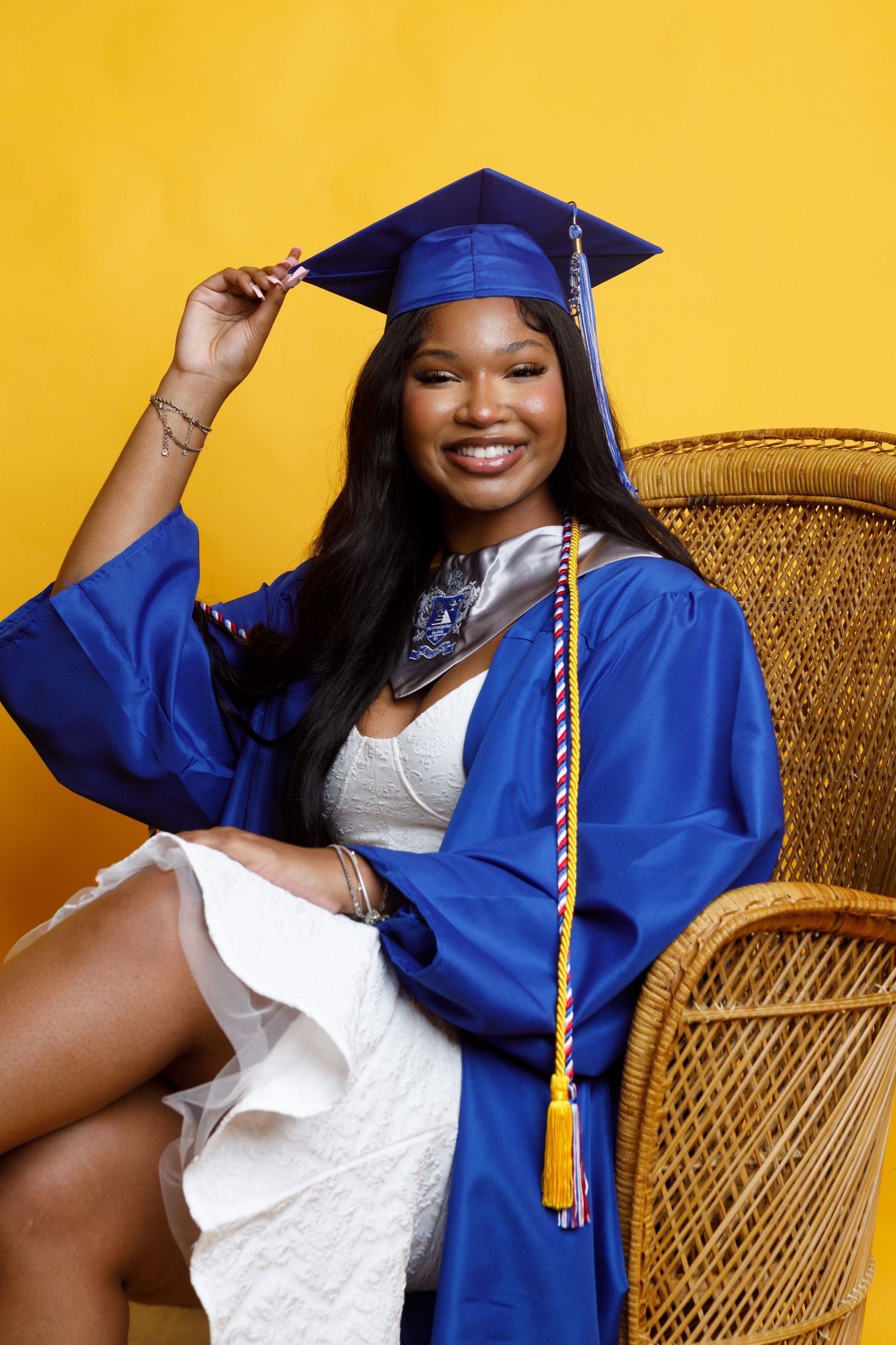 Smiling young woman in blue graduation cap and gown seated in woven chair.