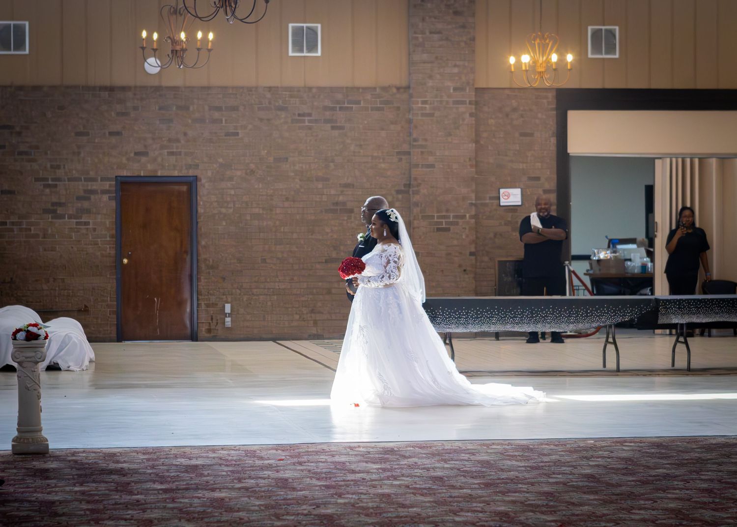 Bride and father entry in elegant wedding reception hall with brick walls and chandelier lighting.