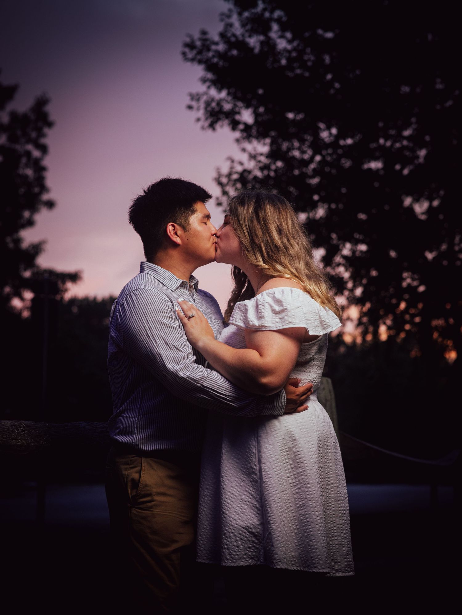 Couple sharing a romantic kiss at sunset with trees silhouetted against a purple and pink sky.