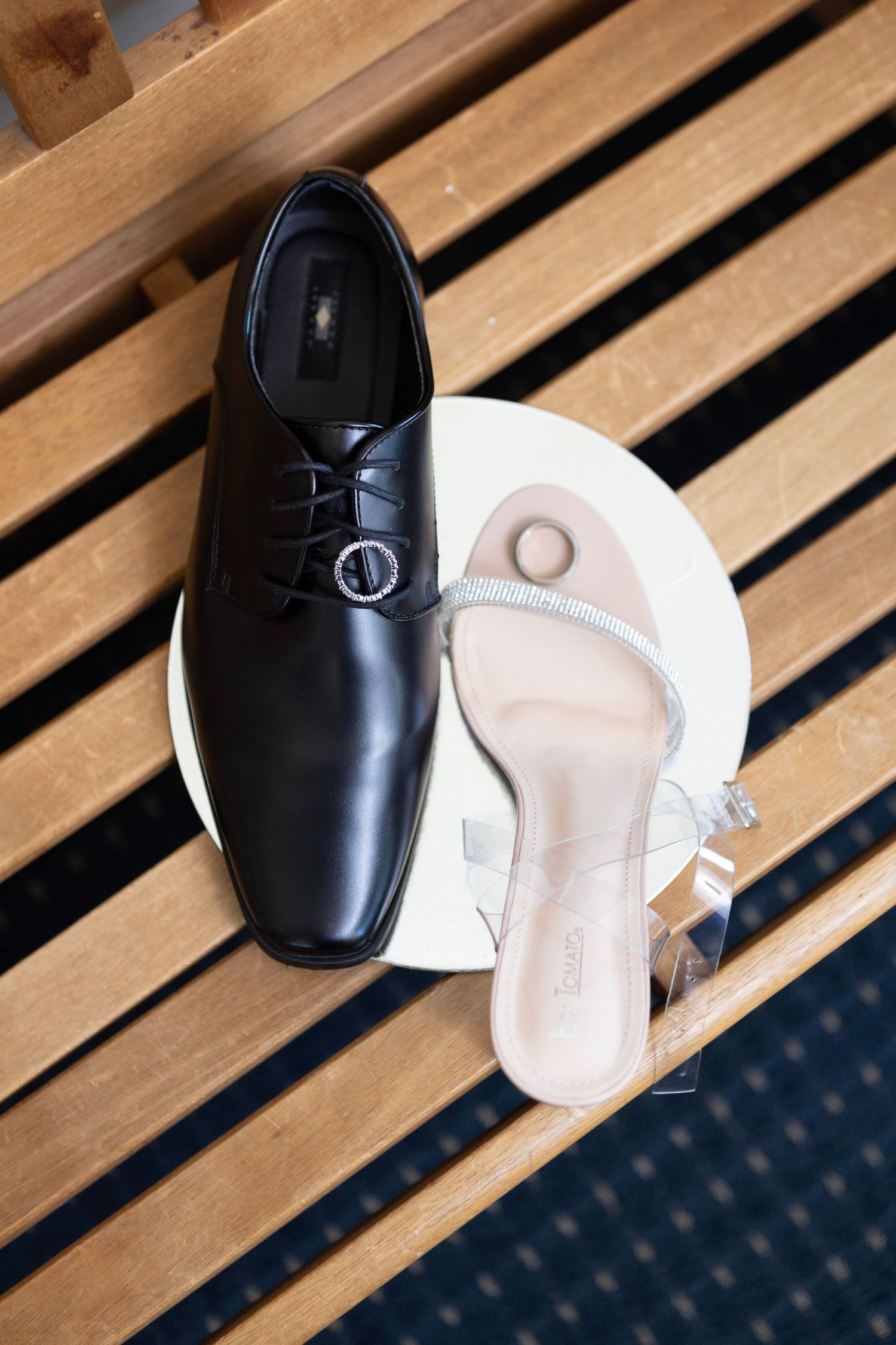 Black leather dress shoe and clear strappy sandal displayed on white plate atop wooden bench for a wedding.