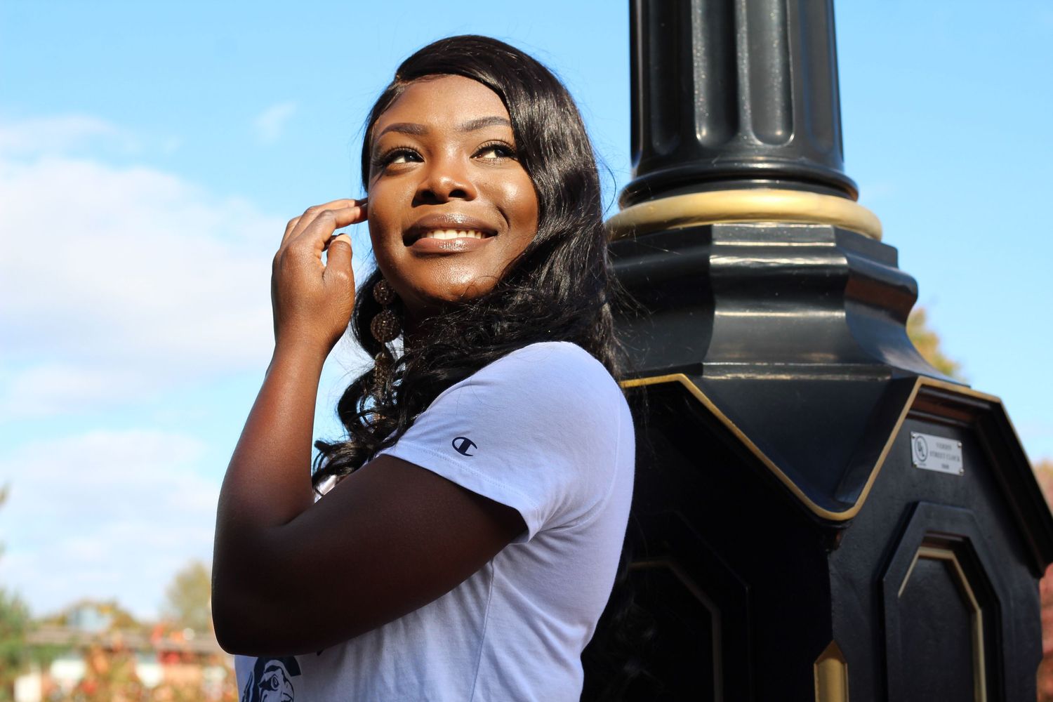 Smiling educated woman in white Champion shirt posing near decorative street lamp outdoors.