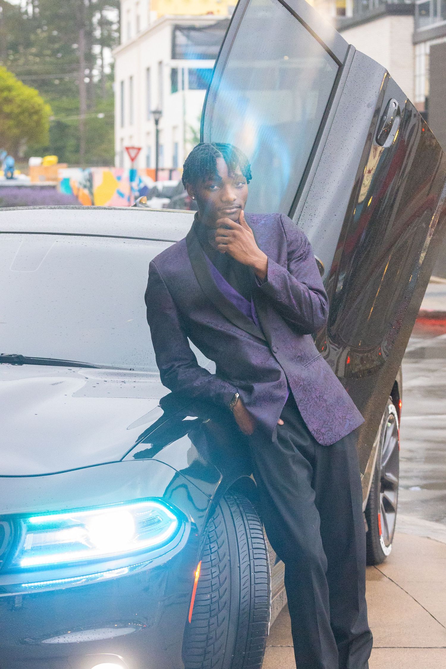Young man in purple blazer leaning against black car with open door for prom.