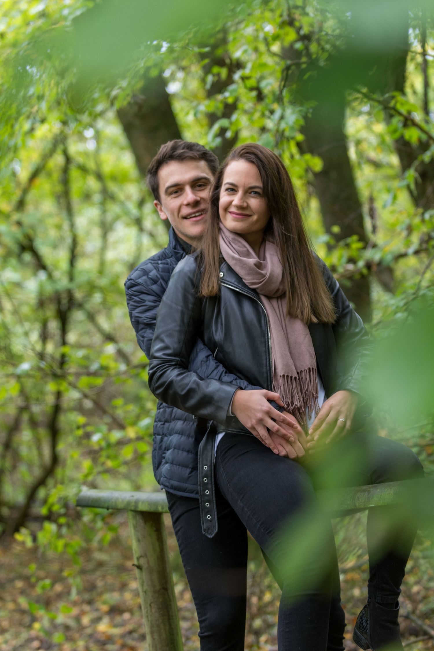 A young couple embracing in a lush green forest, smiling warmly while leaning against a wooden fence.