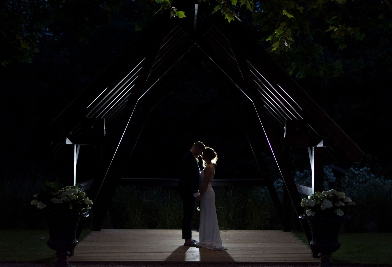 Bride and groom share romantic kiss under illuminated outdoor wedding arch at night.