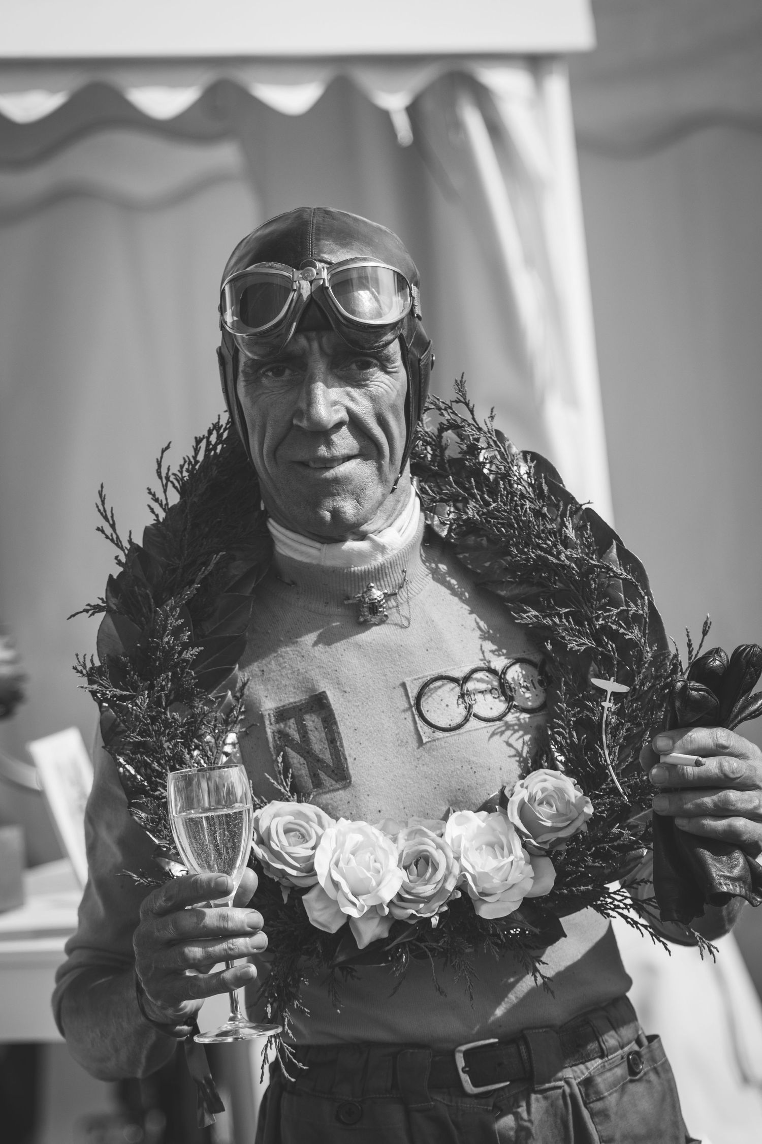 Black and white portrait of a Greame Hardy as racing driver Tazio Nuvolari   wearing goggles and a floral wreath holding champagne.