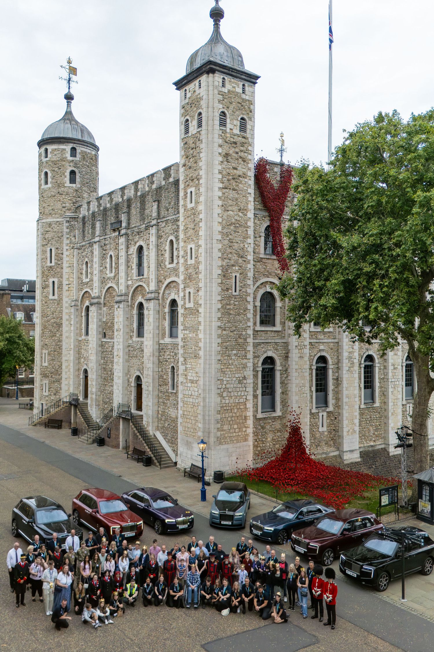 Large group photo outside the Tower of London with luxury cars and a red poppy installation on the historic castle.