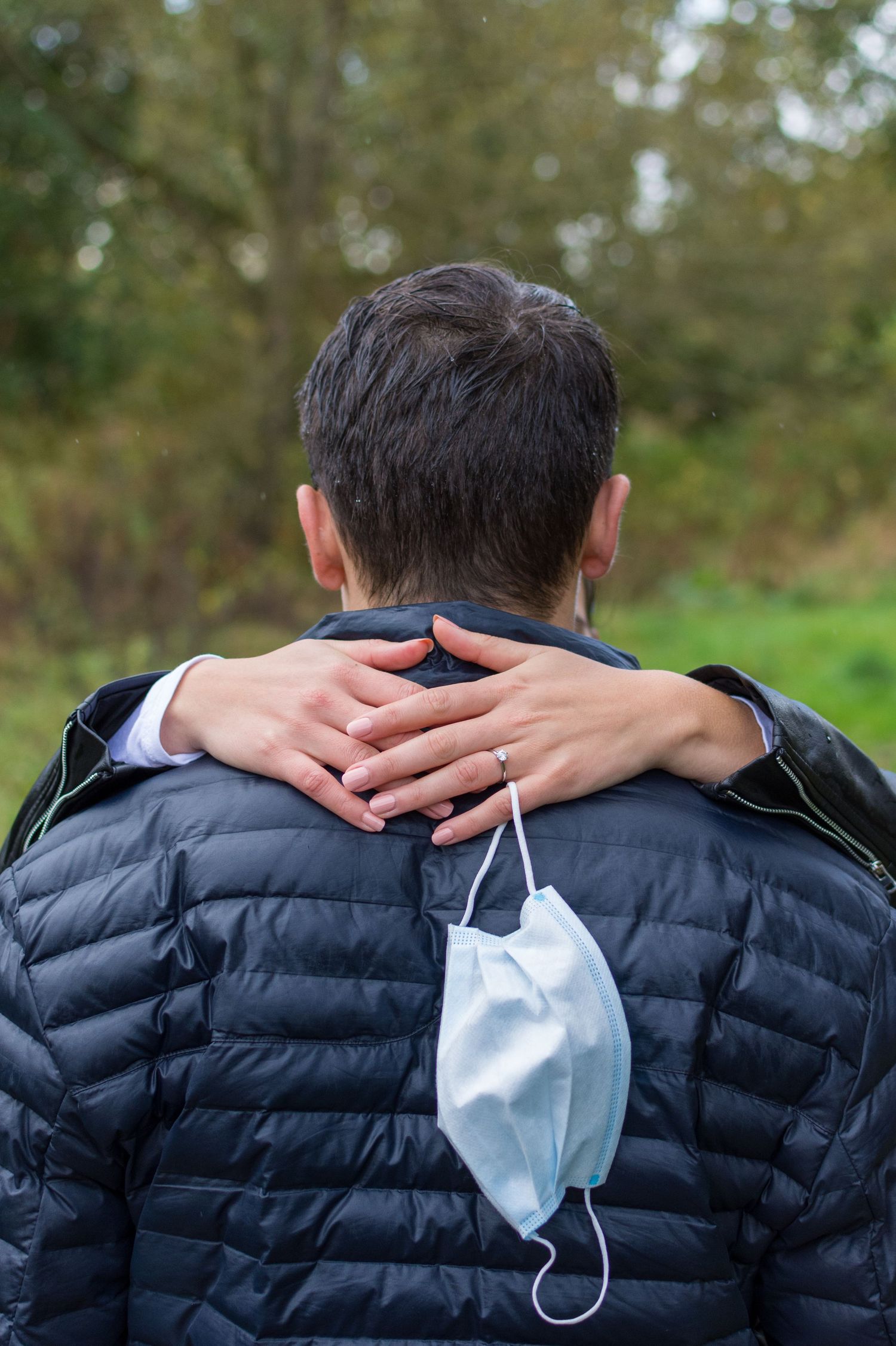Man in black puffer jacket with surgical mask, comforted by a hand on his shoulder outdoors.