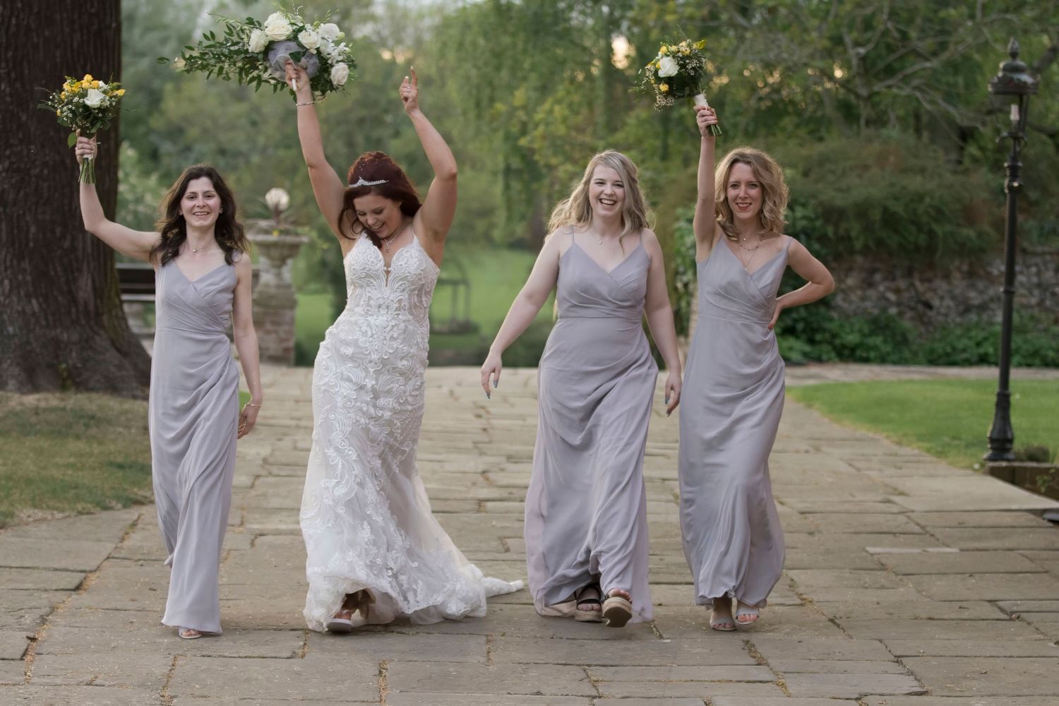 Joyful bride in lace wedding gown celebrates with three bridesmaids in grey dresses holding bouquets on stone pathway.