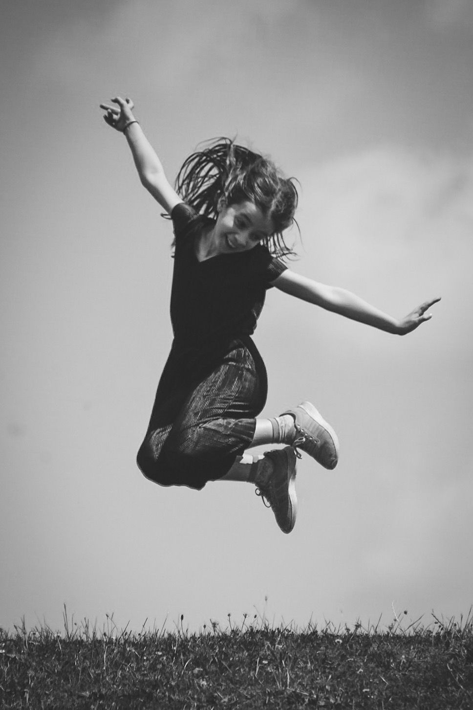 Black and white photo of a joyful young woman jumping high in the air outdoors against a cloudy sky.