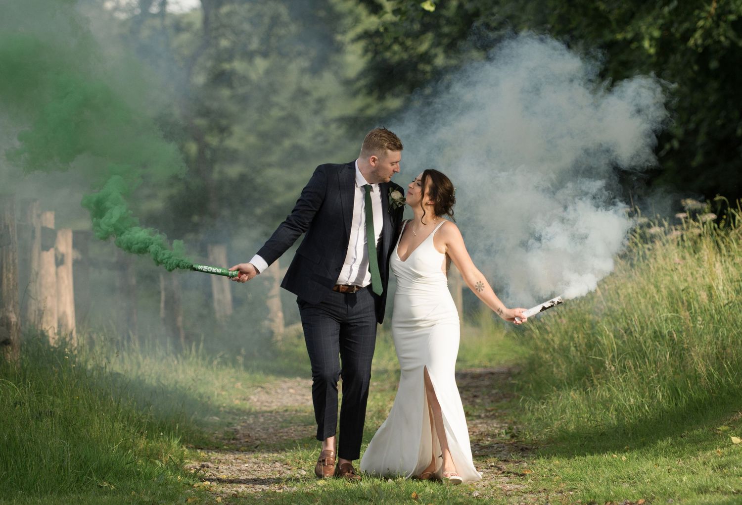 Bride and groom walking outdoors holding green smoke bombs on a scenic woodland path.