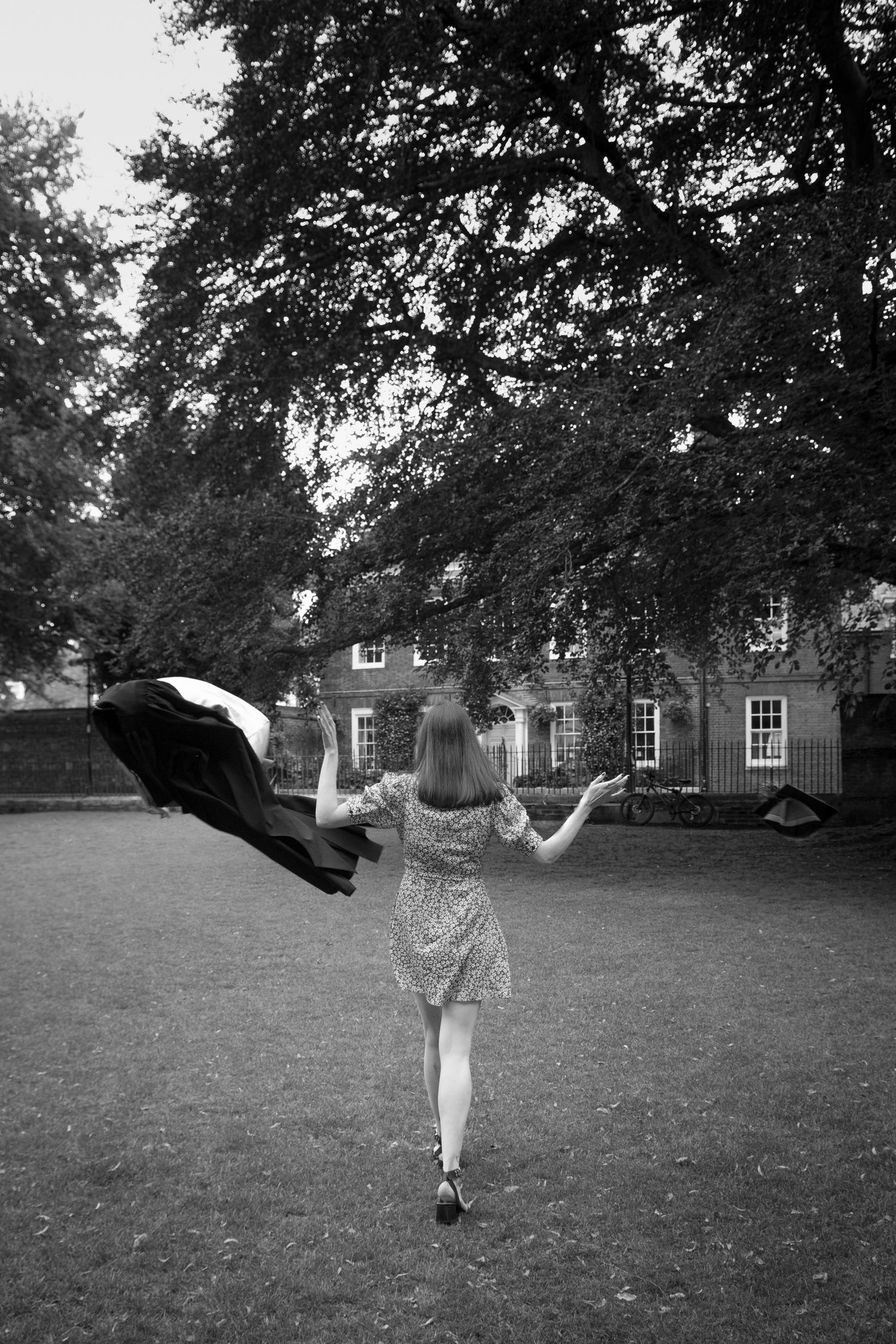 Black and white photo of woman in floral dress tossing her cap & gown in park with large trees after graduating University.