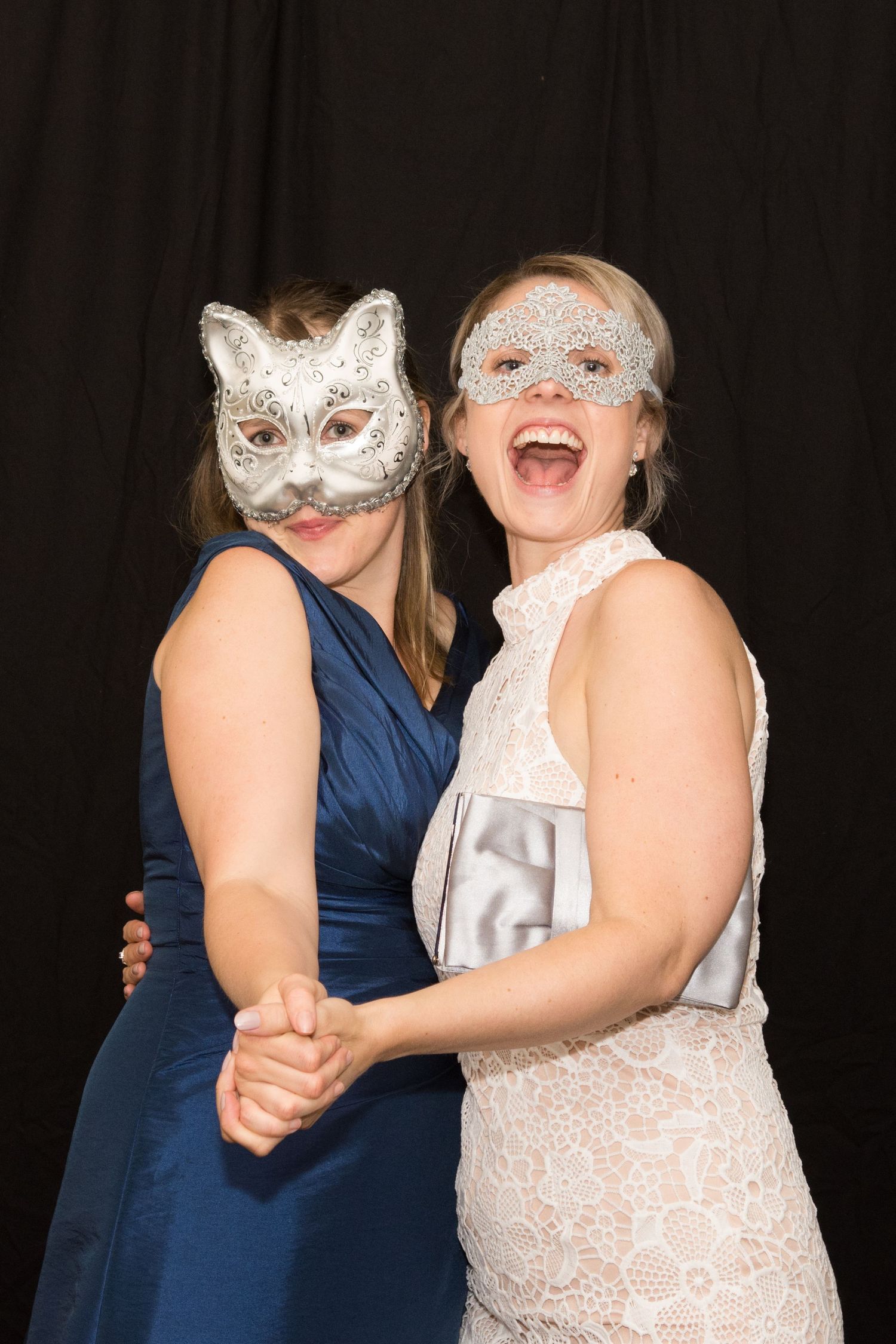 Two women wearing decorative silver masquerade masks smile and pose together against a black backdrop.