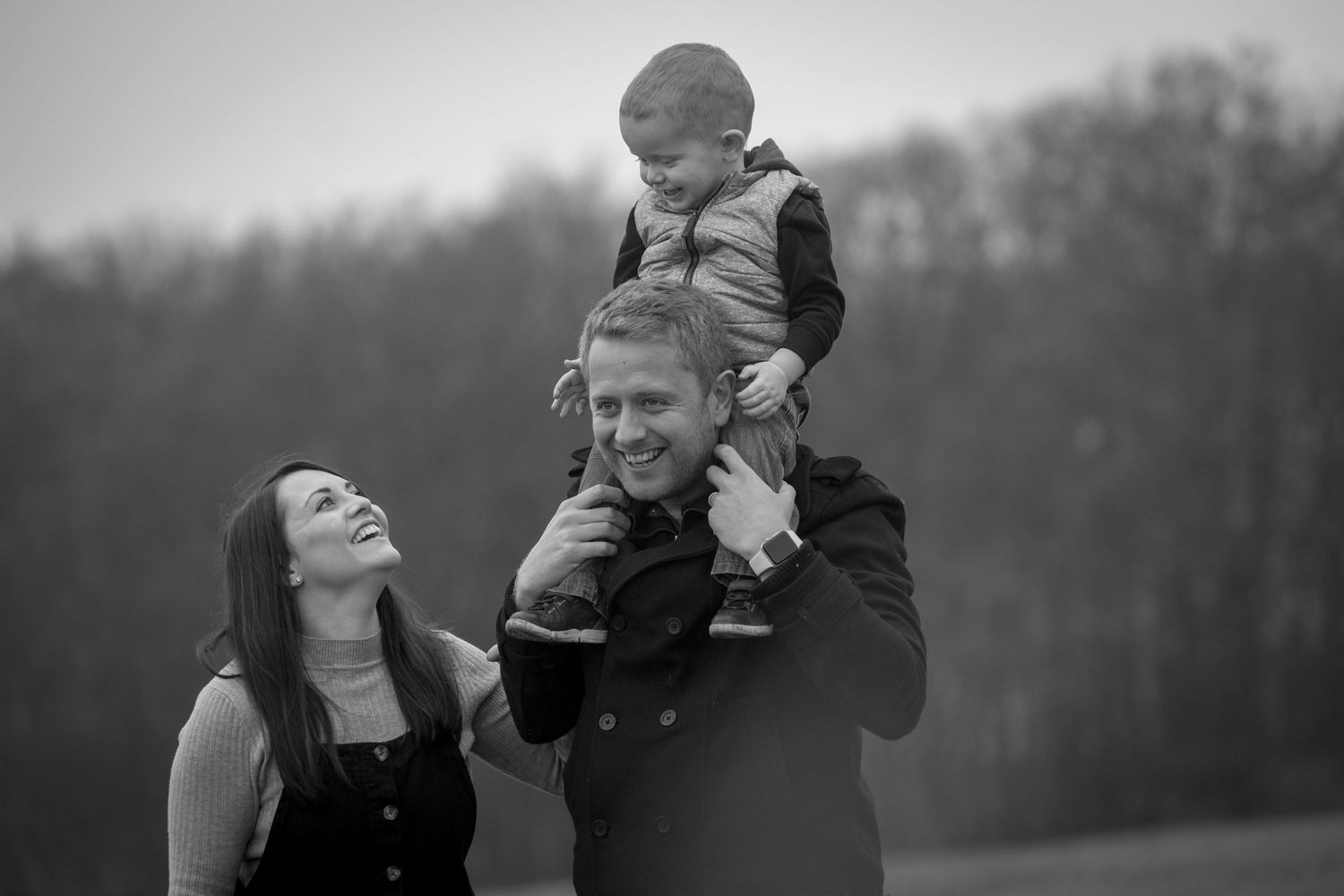 Black and white family portrait of parents laughing with young child sitting on fathers shoulders outdoors.