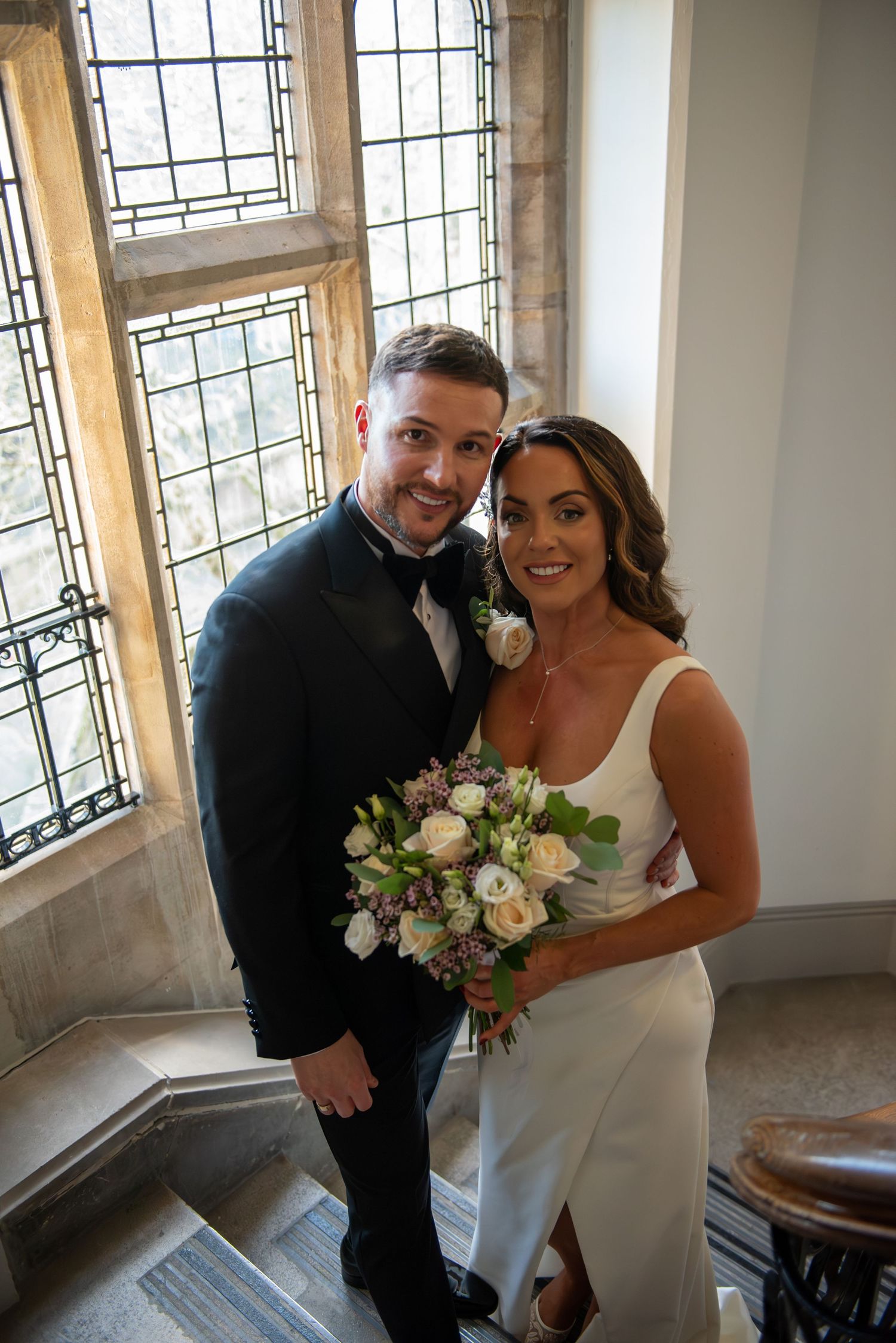 Happy wedding couple posing on staircase by stained glass window, bride holding pastel bouquet.