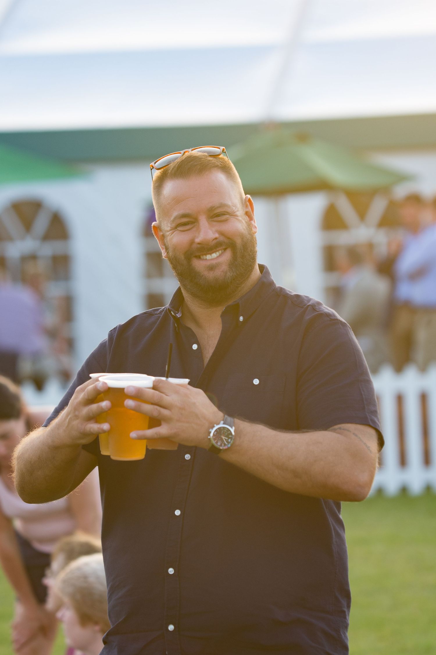 Smiling bearded man holding a large beer at an outdoor summer event with a white marquee tent in the background.