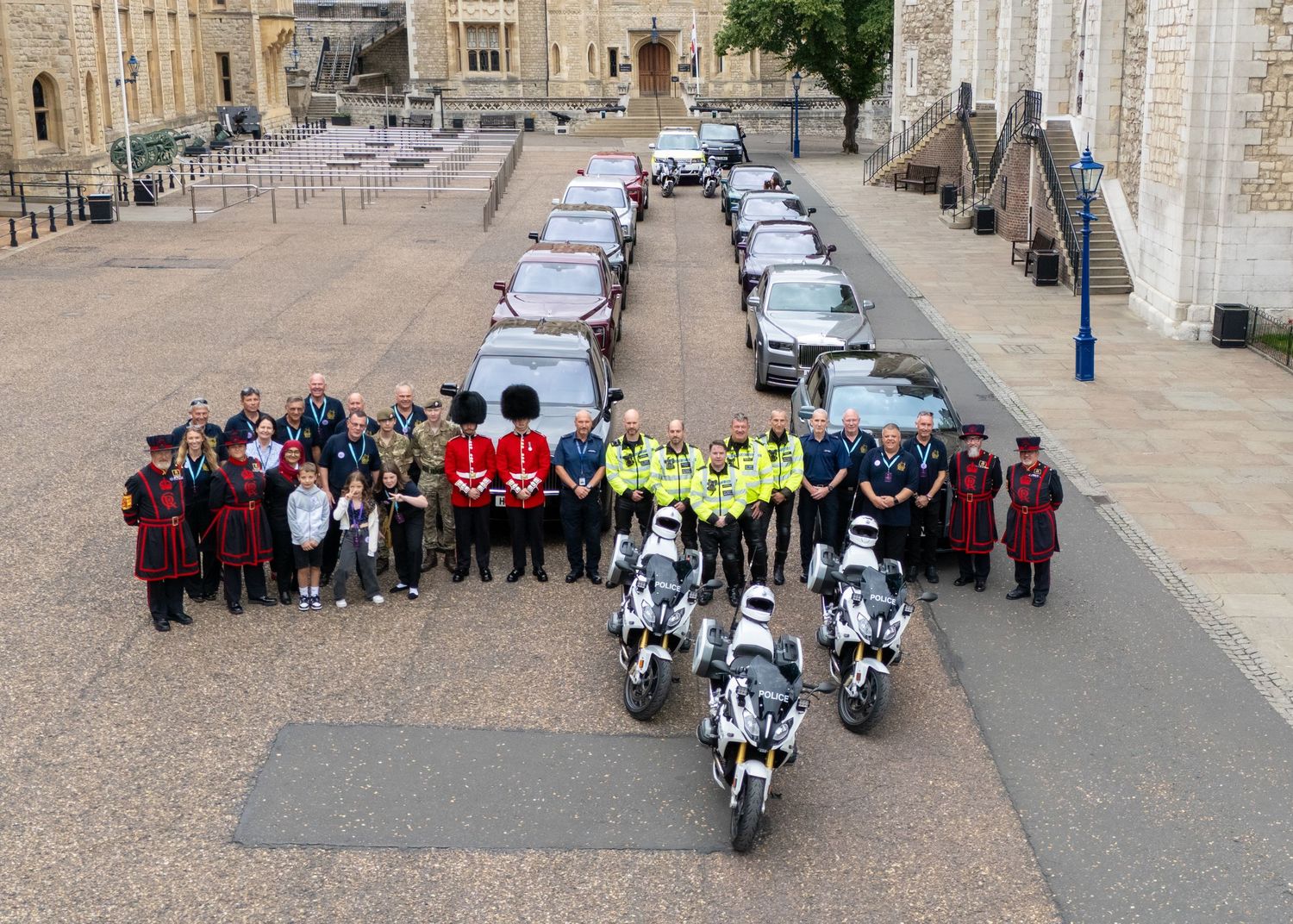 Group of people in uniforms and formal attire posed with motorcycles and cars at the Tower of London courtyard.