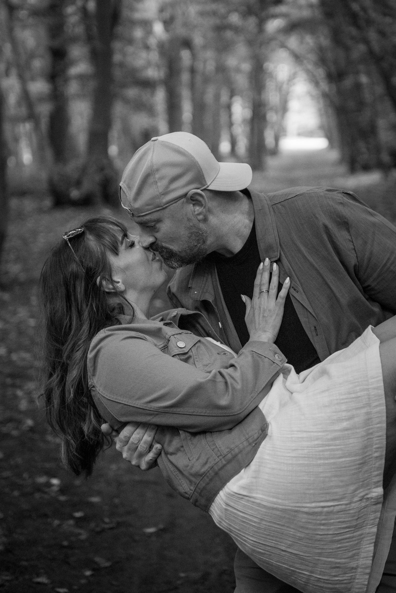Black and white photo of a couple sharing a romantic kiss in a tree-lined park path, woman wearing a denim jacket.