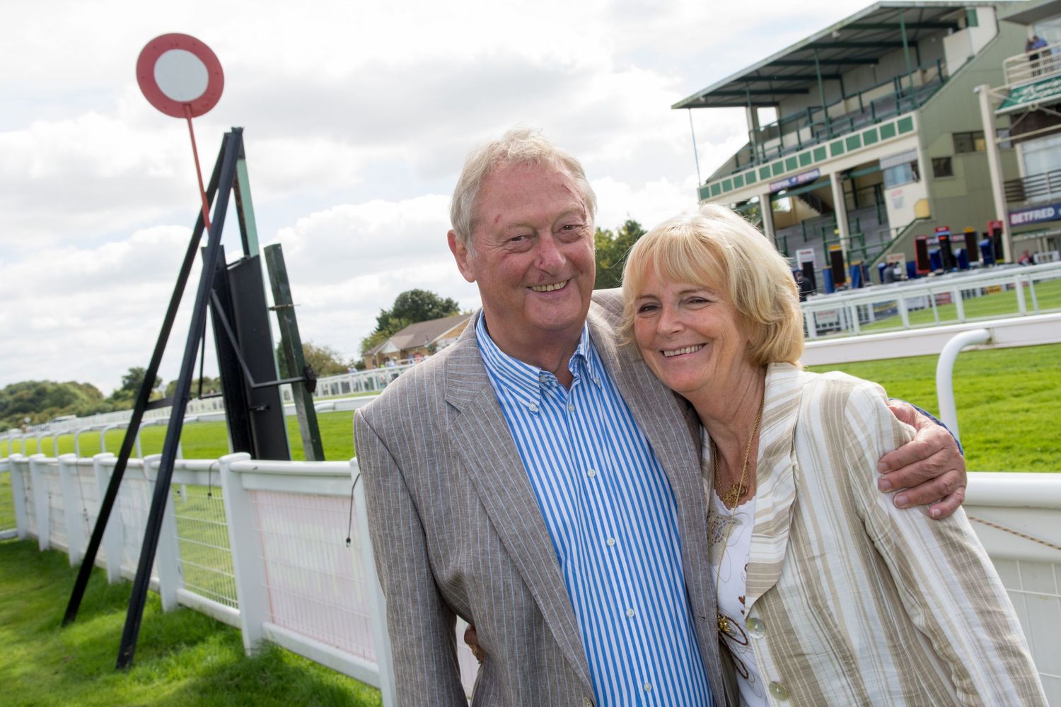 Smiling couple posing together at a horse racing venue with grandstand in the background on a cloudy day.