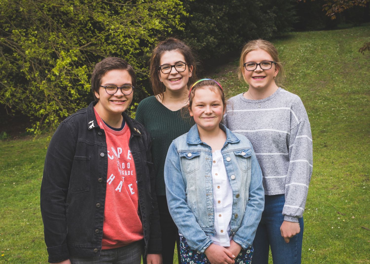 Four smiling women posing together outdoors on a green grassy hill surrounded by trees.
