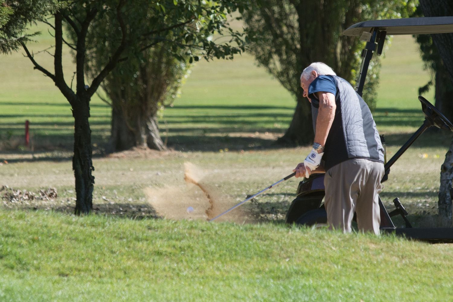 Golfer hitting a sand bunker shot on a lush green golf course surrounded by trees.