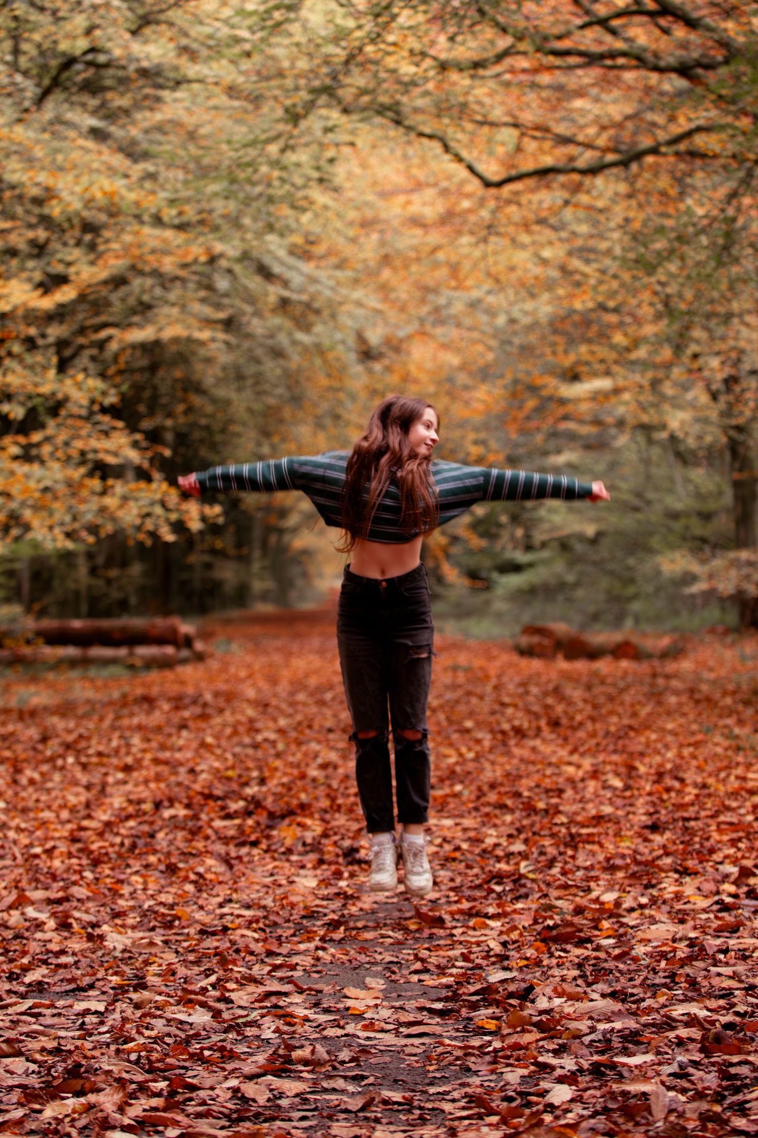 A joyful young woman jumps with arms outstretched in an autumn forest covered in vibrant orange and red fallen leaves.