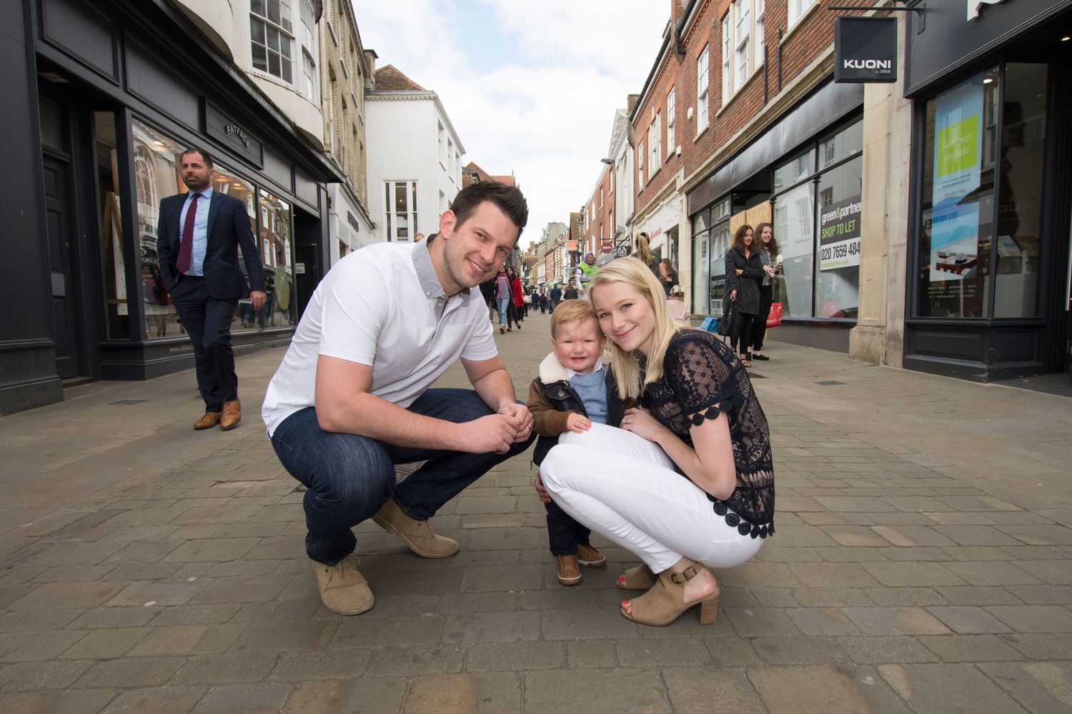 Family of three posing on a pedestrian shopping street, with parents crouching beside young child in a busy town center.