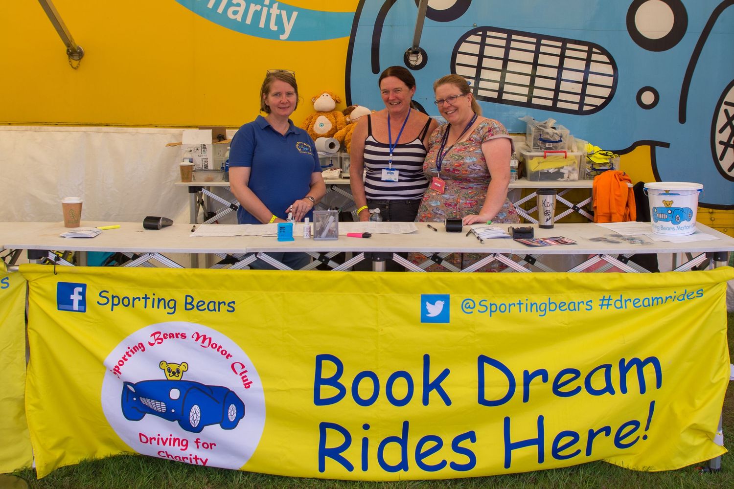 Three women smile at a Sporting Bears charity booth with a yellow banner reading Book Dream Rides Here at The British Motorshow