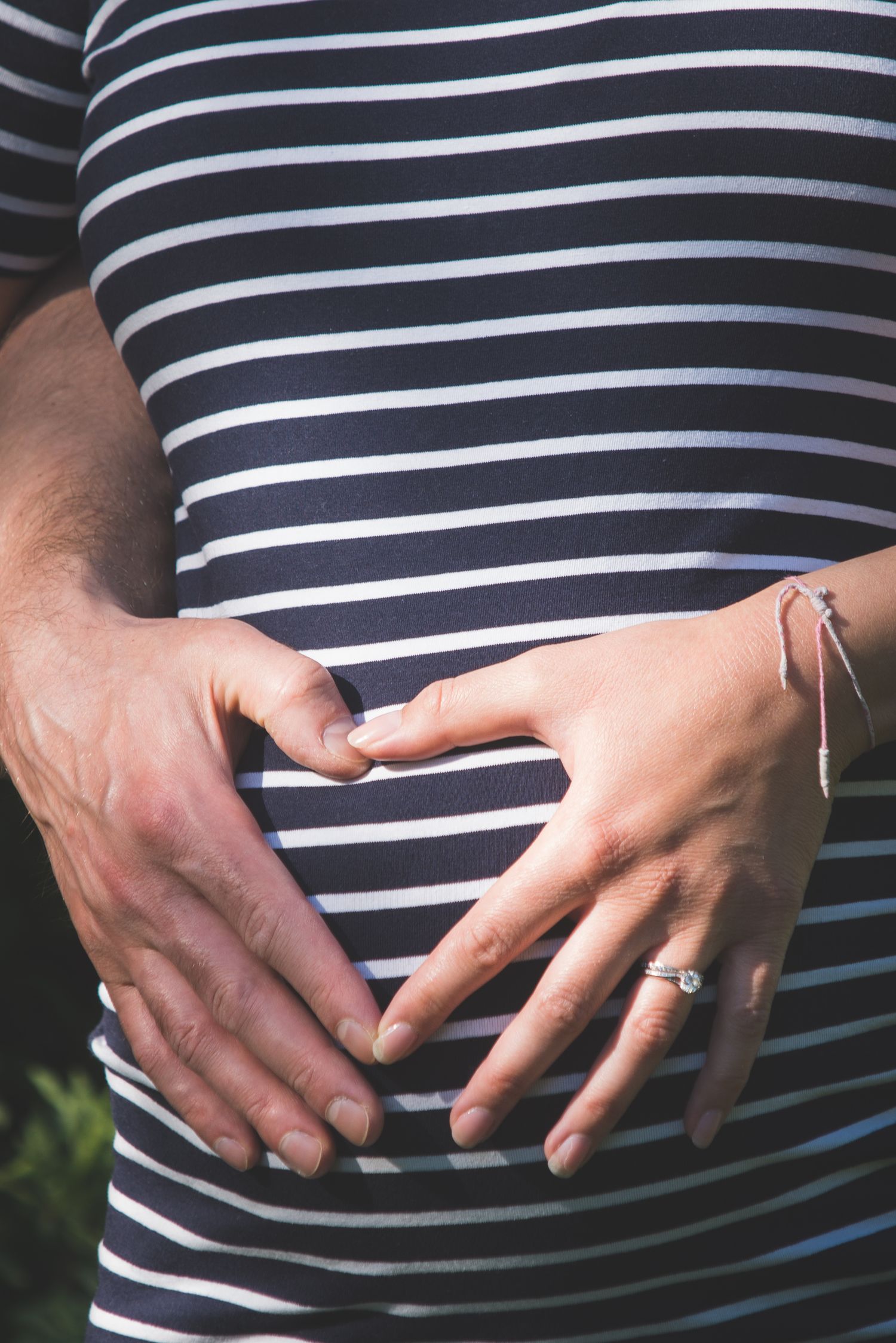 Couple tenderly cradling pregnant belly with hands forming heart shape, wearing striped shirt and diamond engagement ring.