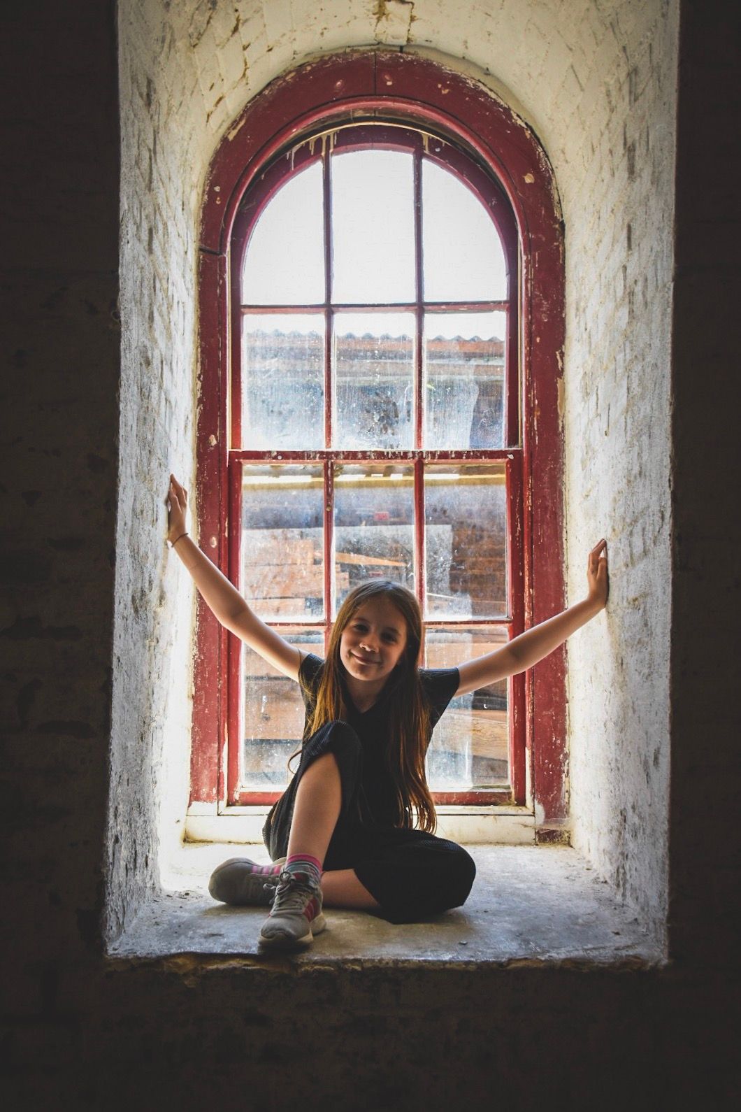 A smiling girl sits cross-legged on a windowsill with arms outstretched in an arched red-framed window of a rustic building.