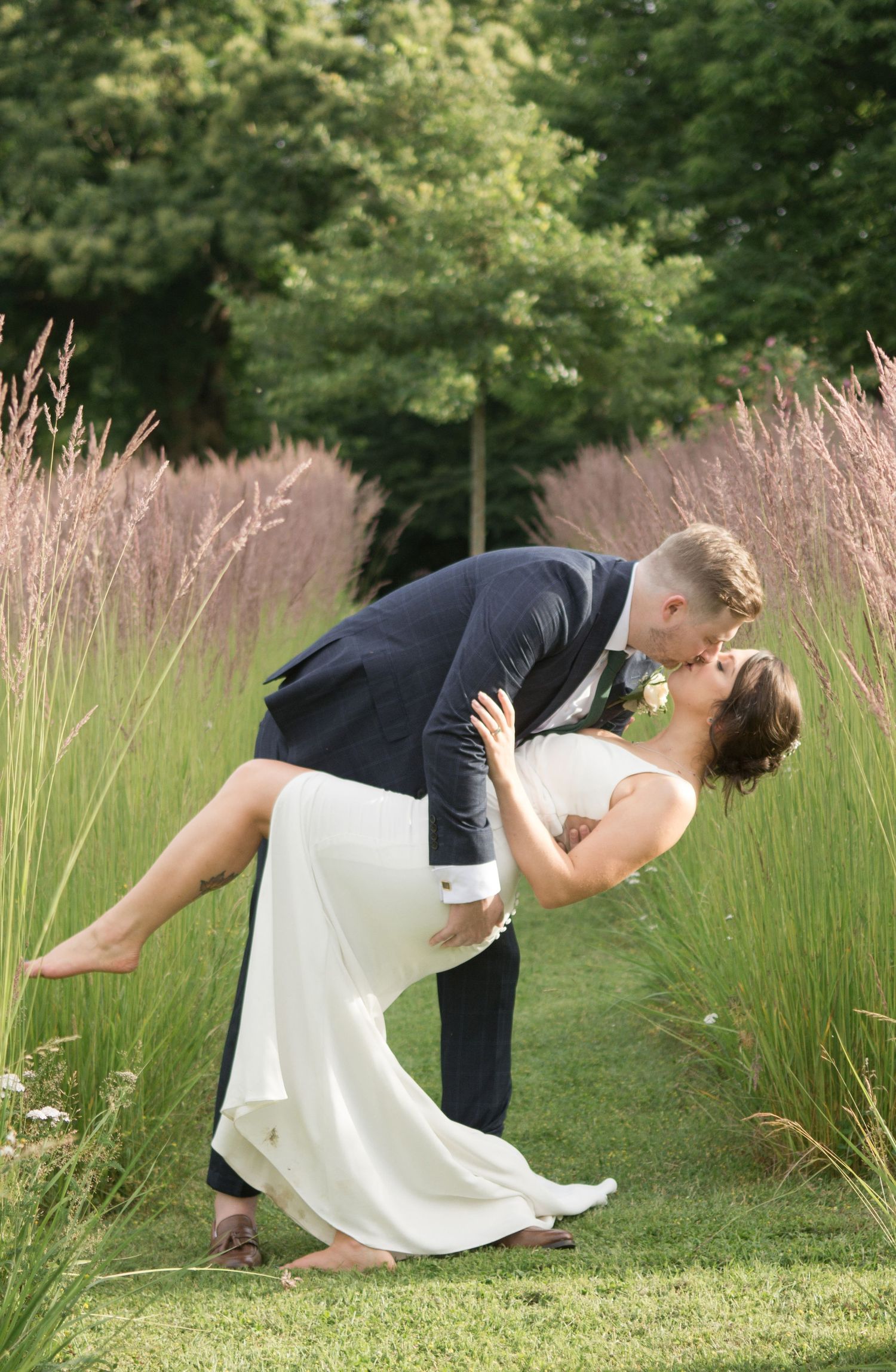 Groom dipping bride for a romantic kiss among tall ornamental grasses in garden setting.