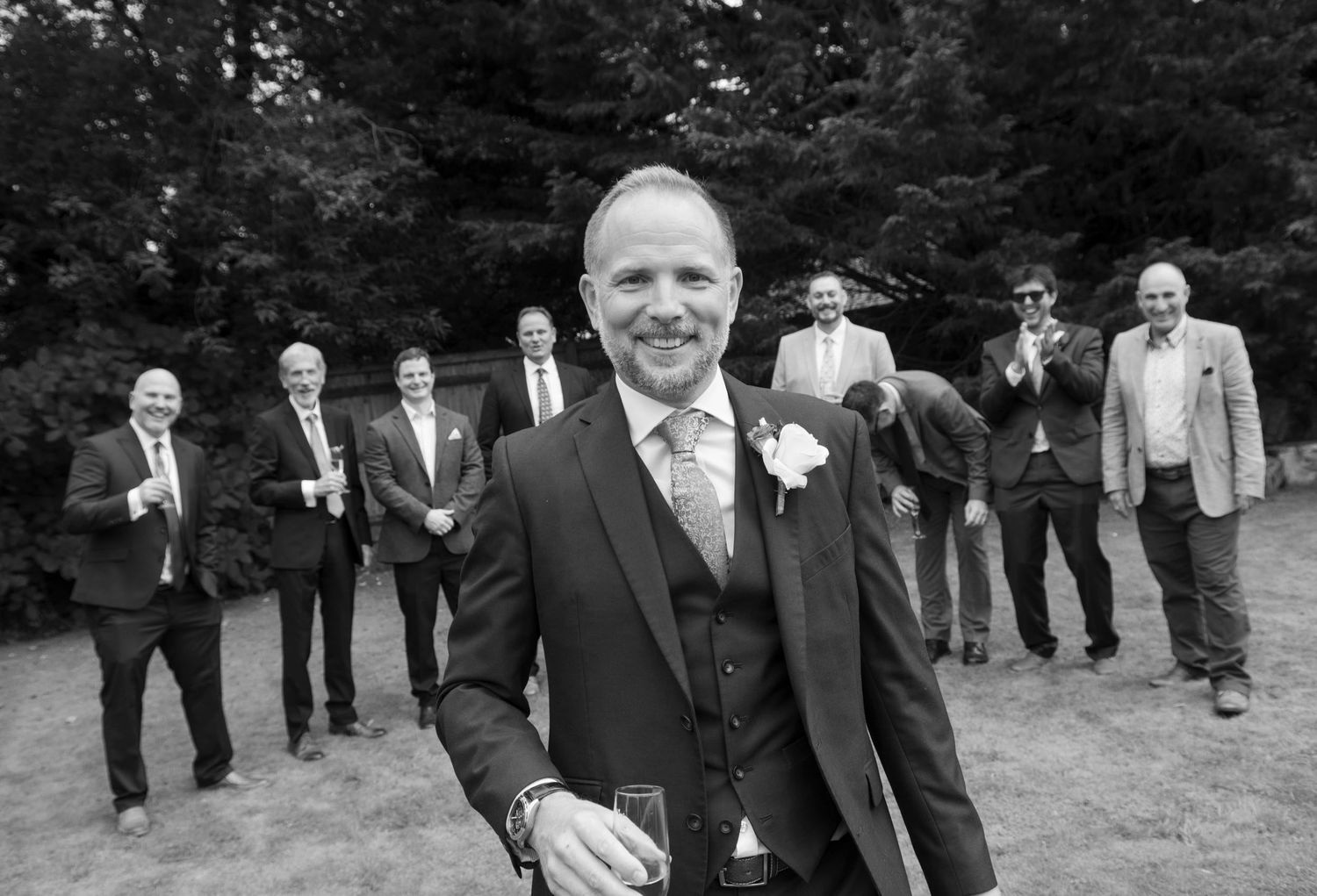 Smiling groom poses with groomsmen in black and white wedding party photo outdoors.