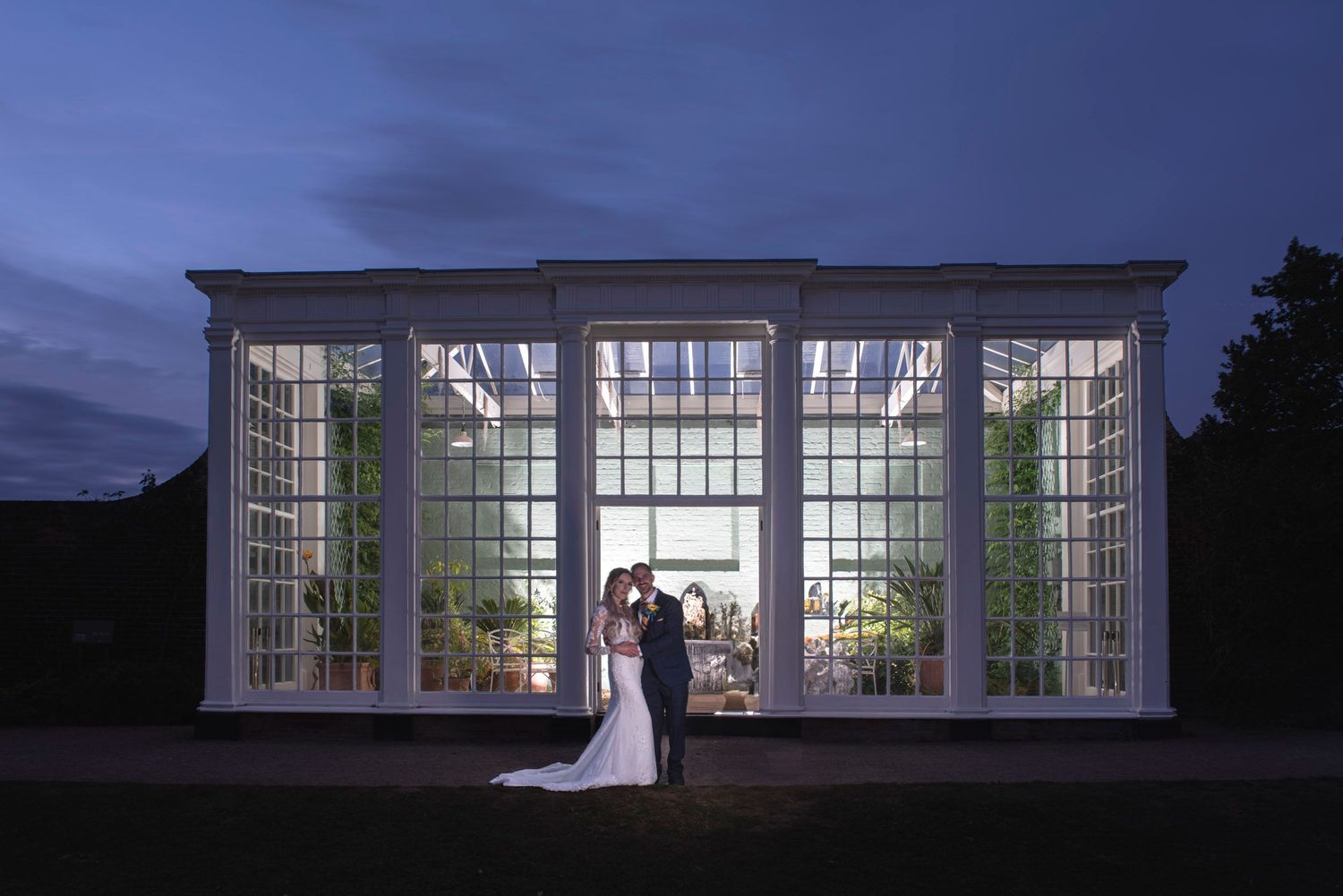 Bride and groom silhouetted against an illuminated glass greenhouse at twilight during wedding.