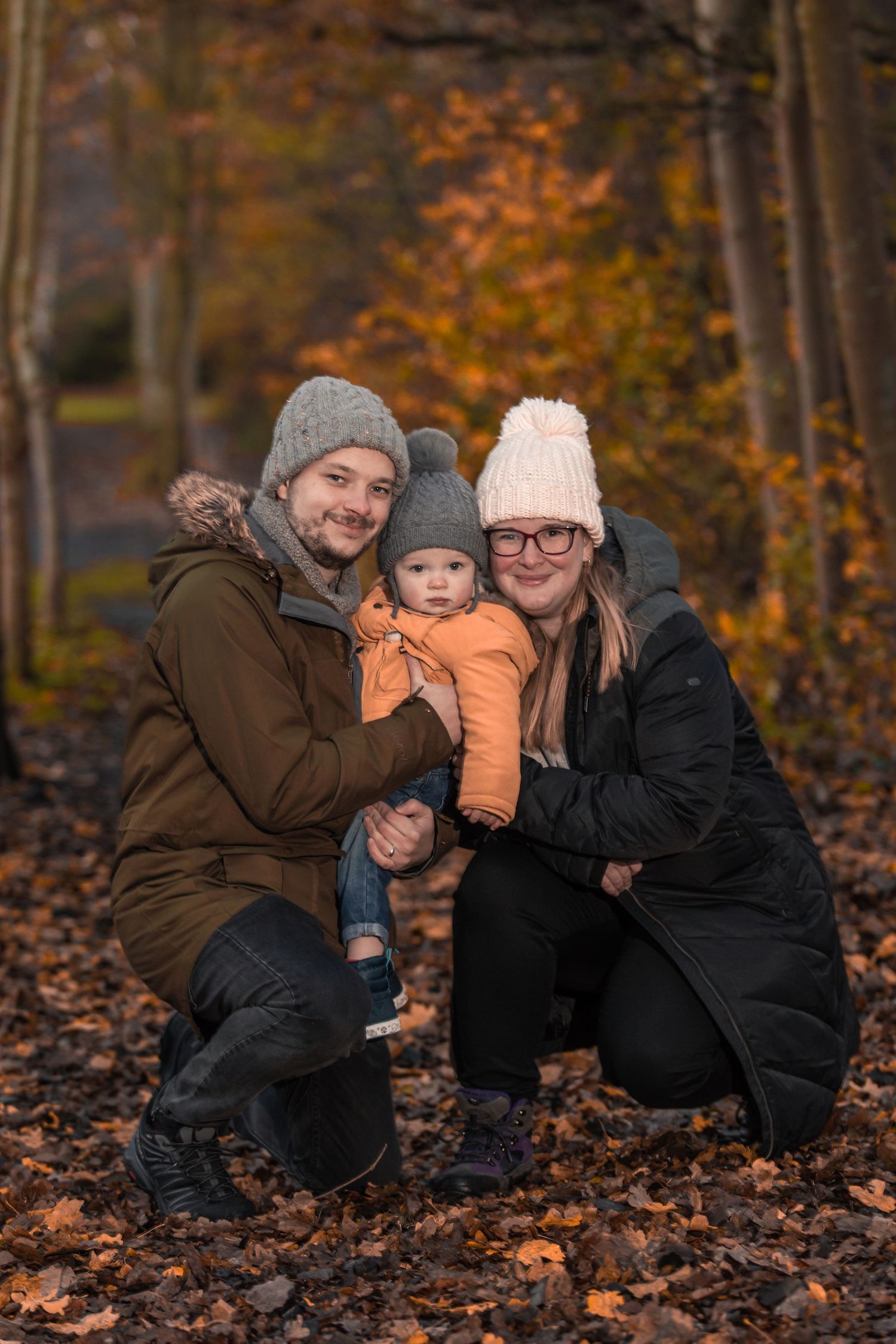 Family of three posing in autumn forest, parents in winter coats and hats crouching with smiling baby in orange jacket.