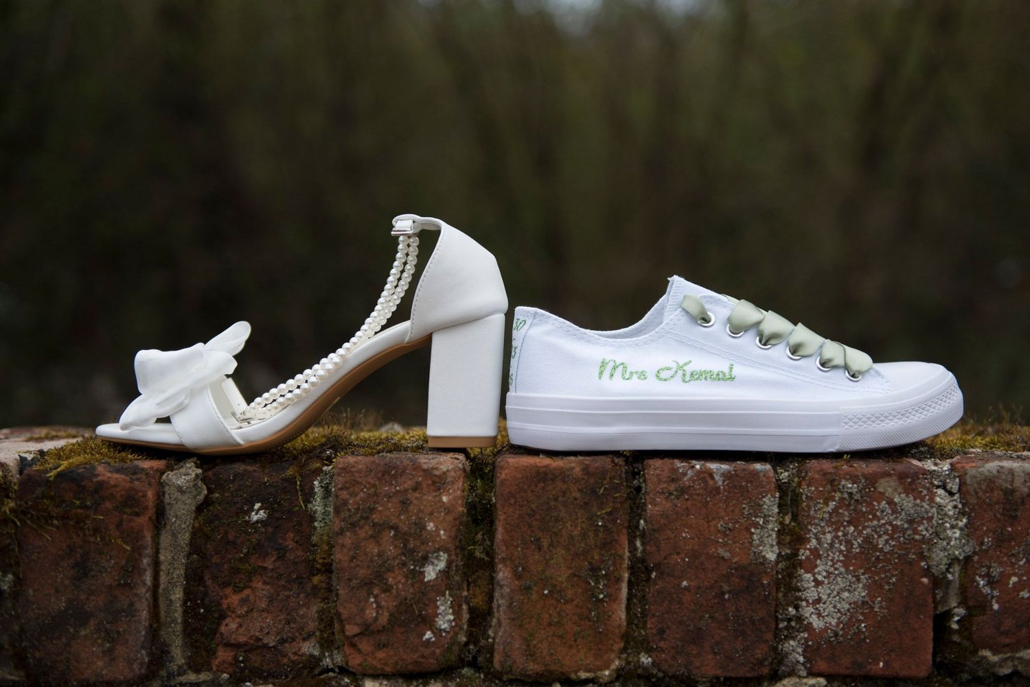 White bridal heeled sandal and personalized white sneaker reading Mrs. Kozak displayed on rustic brick wall outdoors.