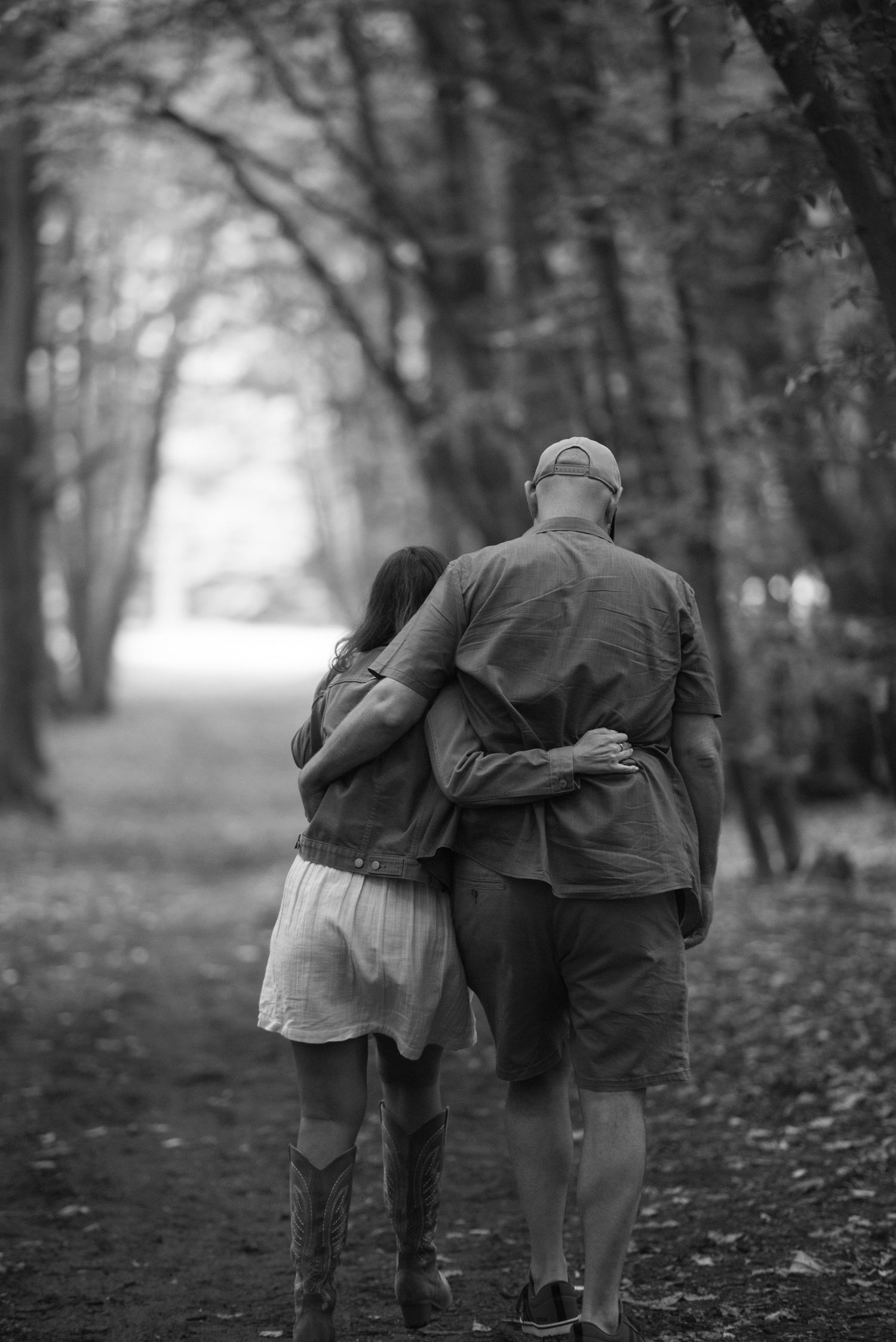 Black and white photo of a couple walking arm in arm down a tree-lined path, viewed from behind.