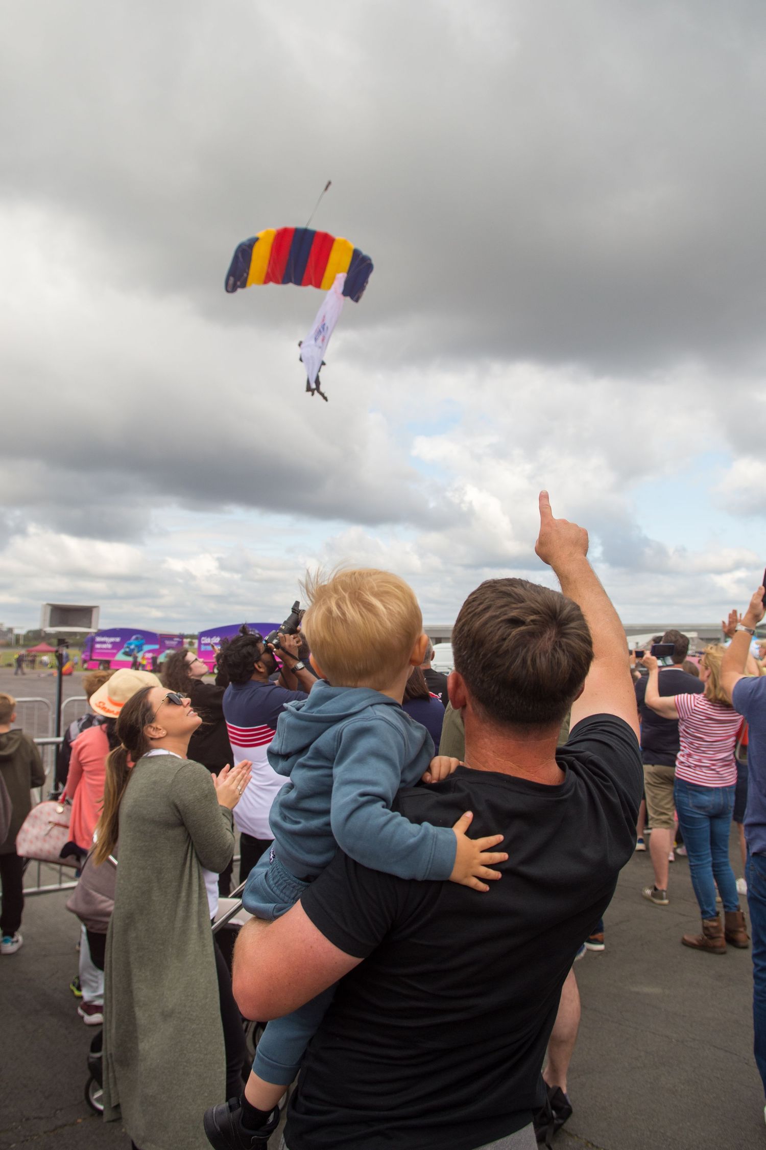 Man holds child pointing at colorful parachutist descending at an airshow, crowd watching under cloudy skies.