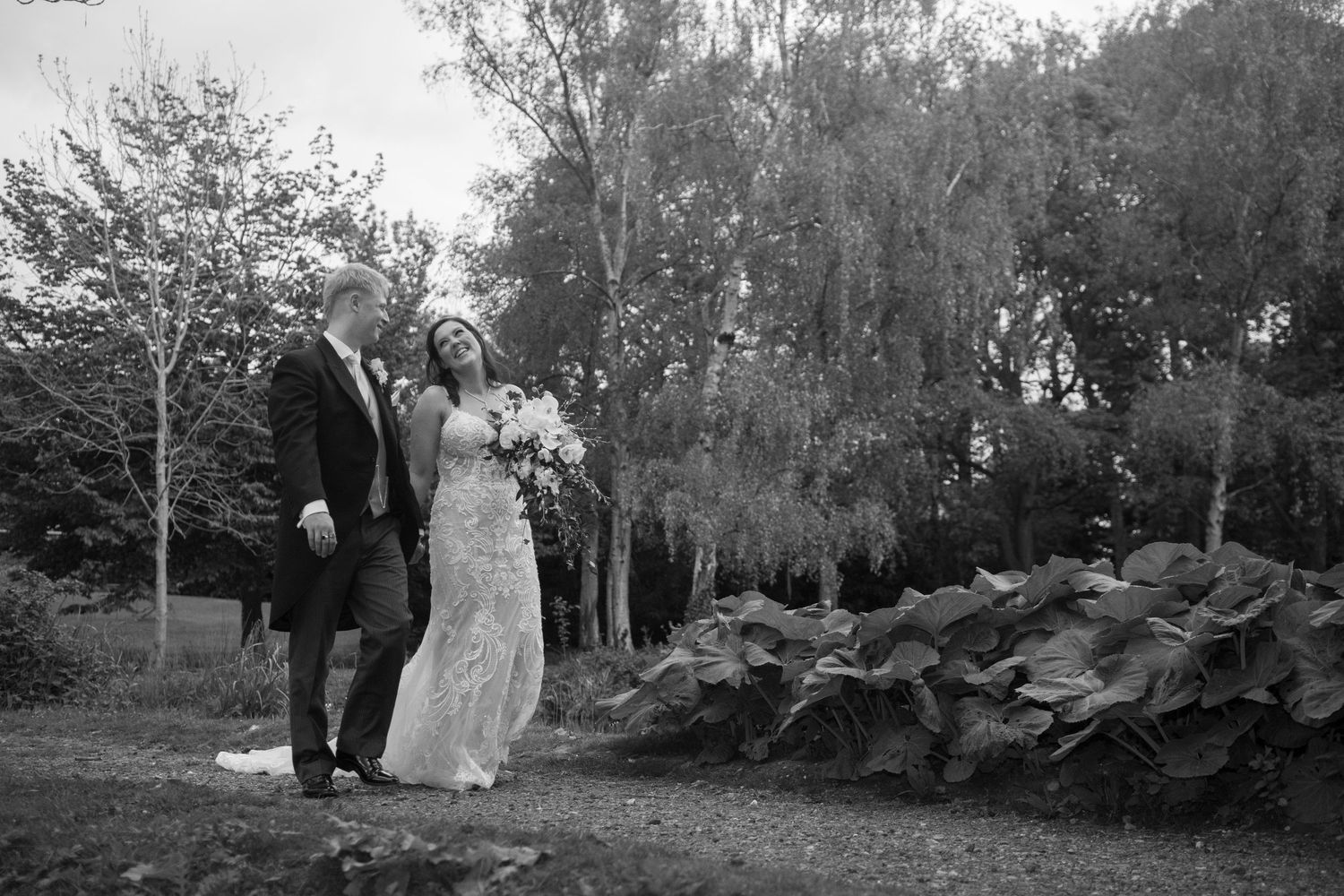 Black and white photo of bride in fitted lace gown and groom strolling through lush garden landscape.