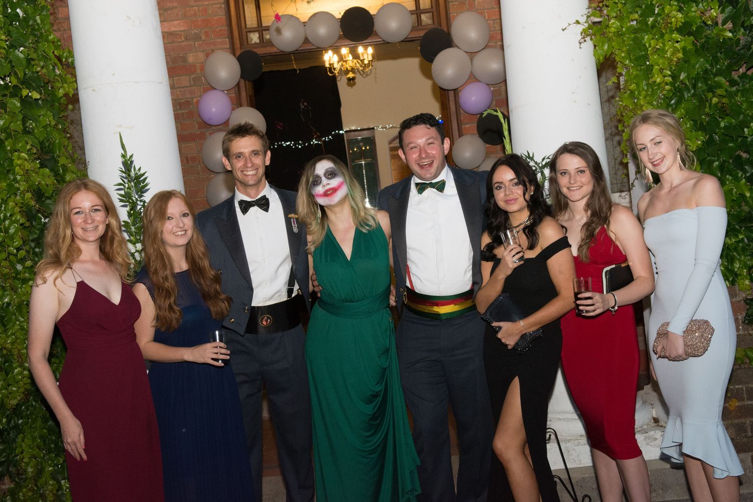 A group of eight elegantly dressed people pose together at a formal masquerade ball event indoors.