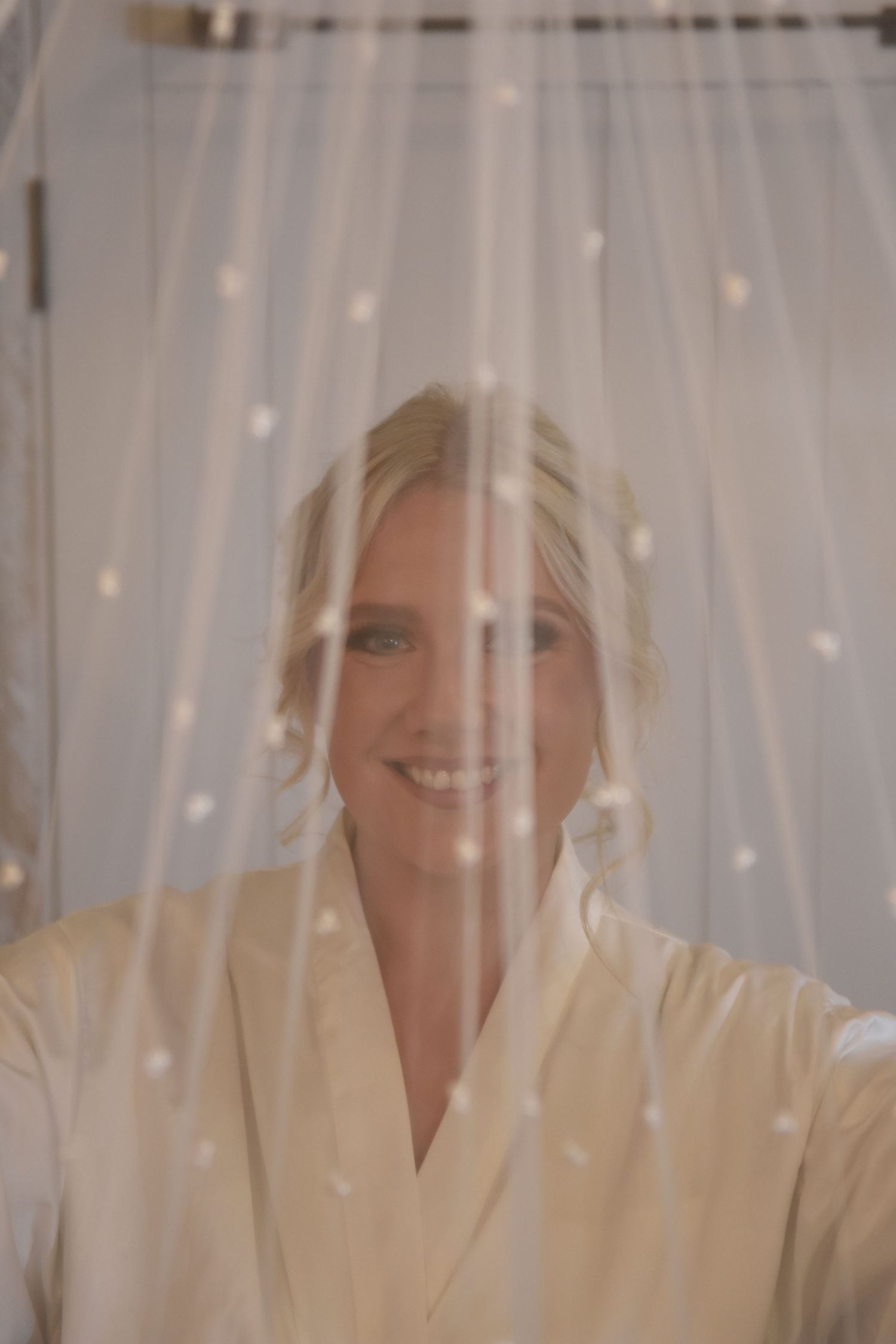 Smiling blonde bride in white robe seen through a delicate pearl-dotted wedding veil.