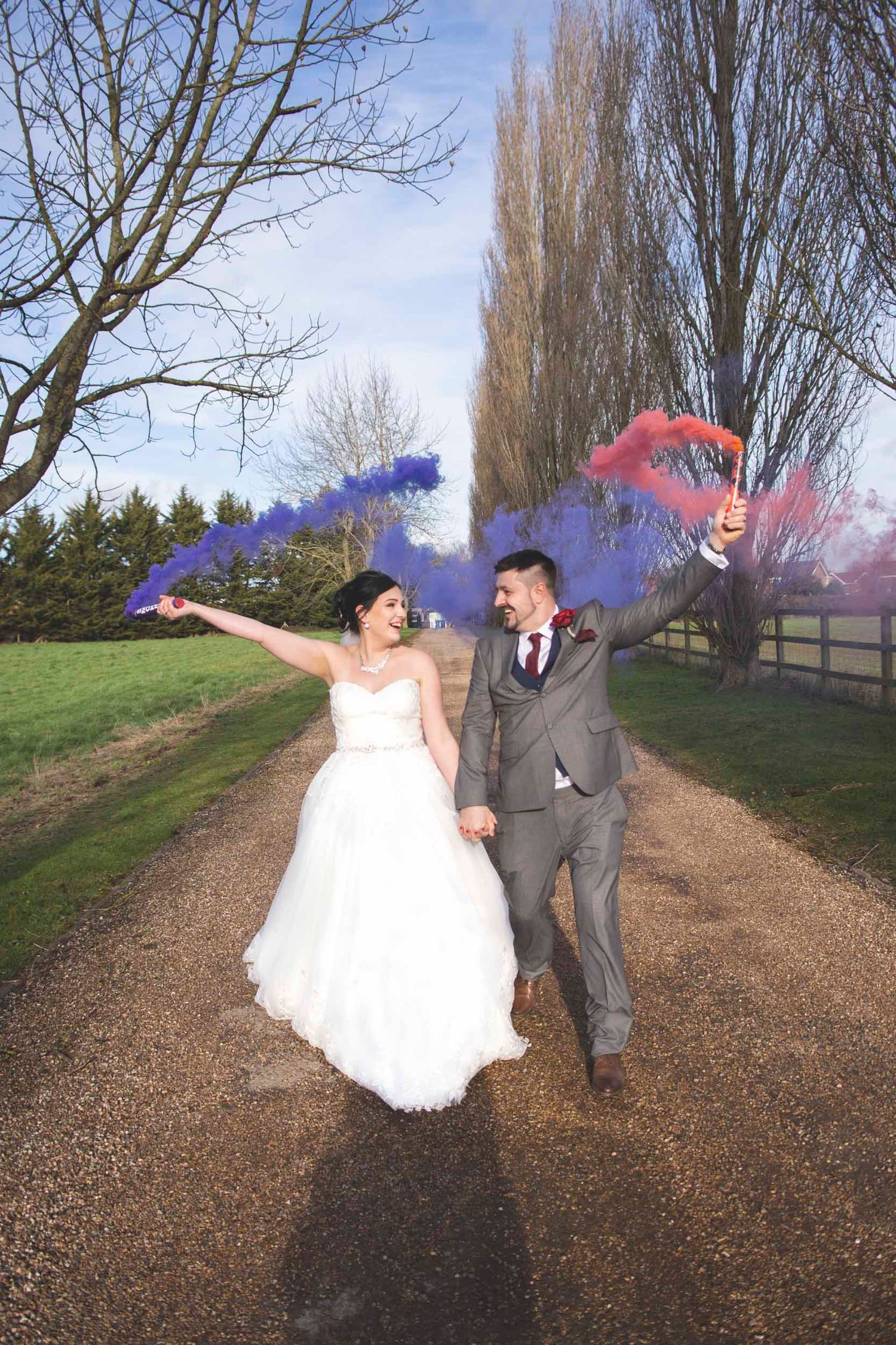 Joyful newlyweds holding colorful blue and red smoke bombs while walking hand in hand down a tree-lined gravel path.